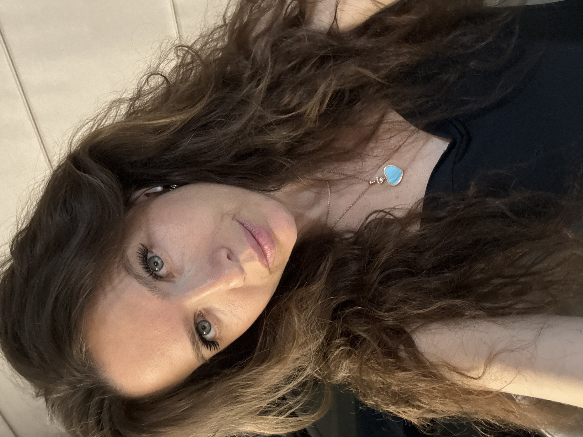 A woman with long, brown, wavy hair and light eyes is the subject of this close-up shot. She is wearing a black top and a delicate necklace with a blue heart-shaped pendant. The background appears to be a light-colored wall with vertical panels. There is no information to suggest this image was taken in Dolgoprudny, Russia.