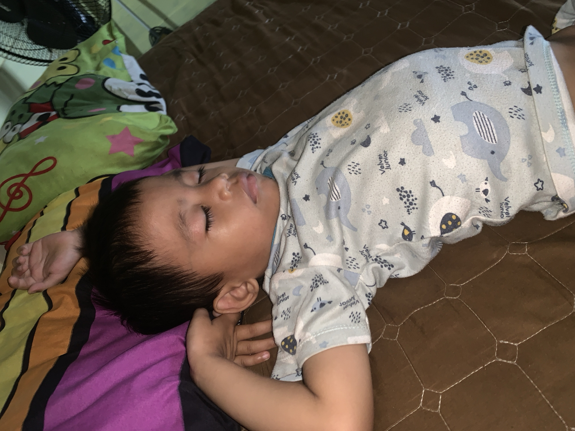 A young child with dark hair is sleeping on their back, with their right arm bent and head resting on their right arm. They are wearing a light blue pajama set with a repeating pattern of cartoon elephants, clouds, and stars. The child is on a brown quilted surface, possibly a mattress or blanket. In the background, there are pillows with colorful striped and cartoonish patterns, and a black electric fan is visible in the upper right corner. The scene appears to be an indoor setting, likely a bedroom. The location is described as Sungai Meriam, Indonesia.