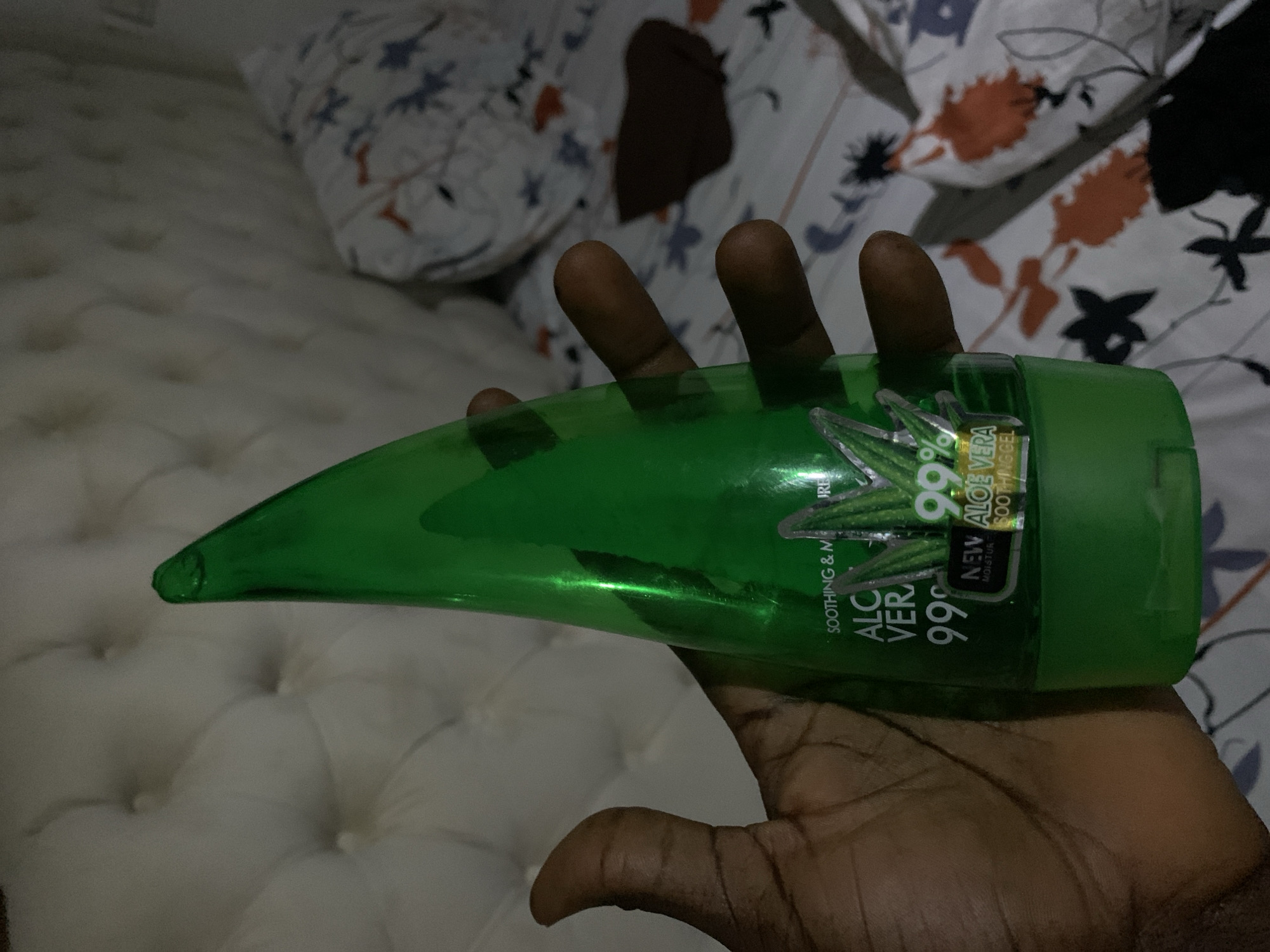 The image displays a dark-skinned hand holding a bright green, leaf-shaped bottle horizontally. The bottle is the main subject and appears to contain an aloe vera product. Visible text on its label includes "99%", "ALOE VERA", "SOOTHING & MOISTURE", "NEW ALOE VERA SOOTHING GEL", "PURE", and "MADE IN MALAYSIA". The label also features green leaf illustrations.
The setting is indoors, likely a bedroom, indicated by the background elements. A white, tufted surface, possibly a mattress or headboard, is visible in the foreground and receding into the background. Behind the hand and bottle, a patterned bedspread or blanket can be seen, decorated with white, orange, grey, and black floral or leaf motifs on a lighter fabric.
The person's hand is holding the bottle steadily, presenting it to the viewer. The lighting in the image is somewhat dim, suggesting an indoor environment with artificial light or low natural light. No specific activity beyond holding the product is discernible. There are no visual cues to indicate the time of day, weather, or emotions. The image is set in an indoor domestic environment, with no visual elements explicitly confirming the Gwarinpa, Nigeria location context.