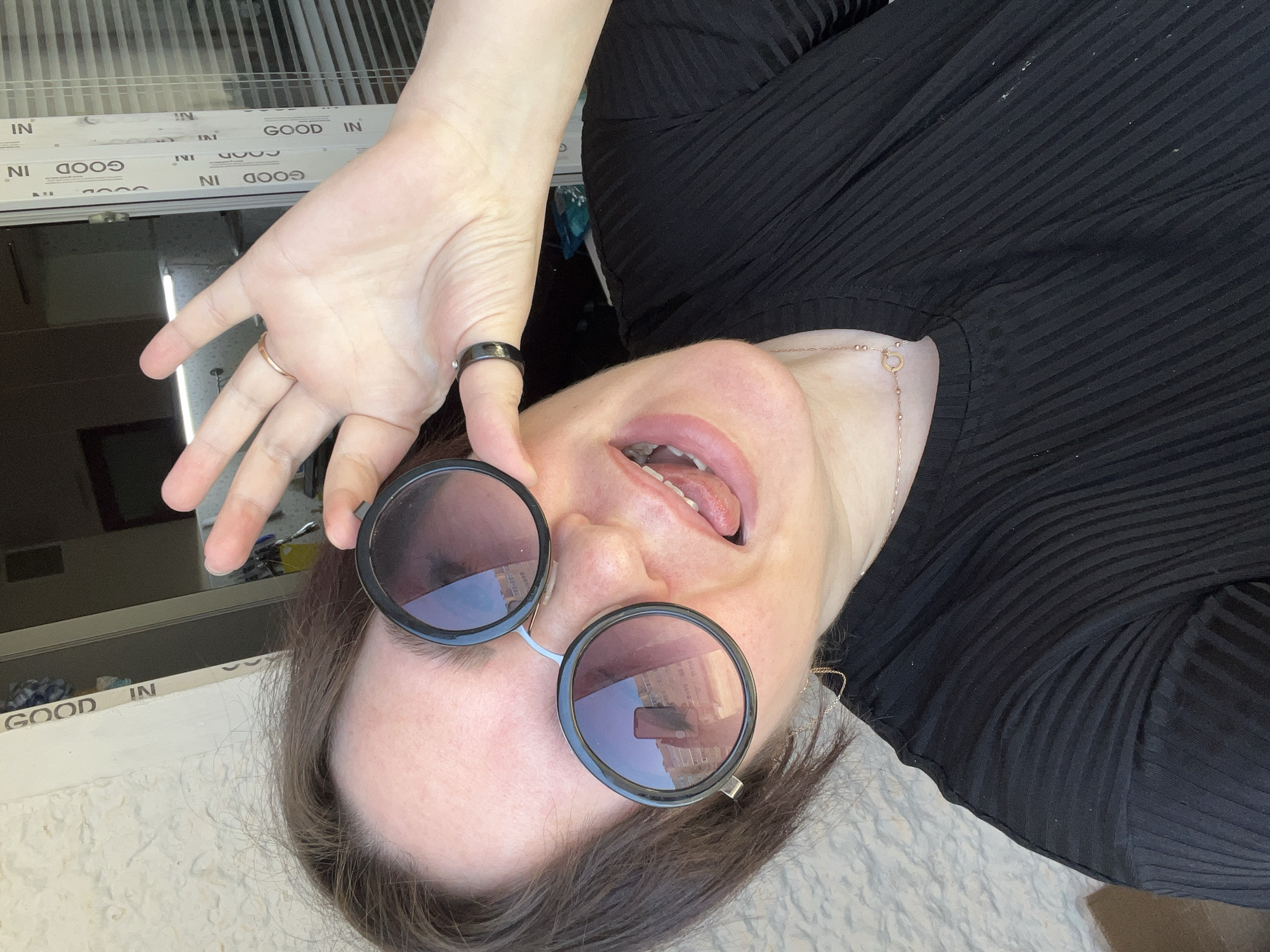 The image depicts a woman indoors, wearing round sunglasses and a black ribbed top. She is sticking her tongue out and holding up her hand, which has two rings on it. The background features a window with "GOOD IN" repeated on the frame. The reflection in the sunglasses suggests a city scene, but the specific location in Noviy, Russia, cannot be determined from this image alone.