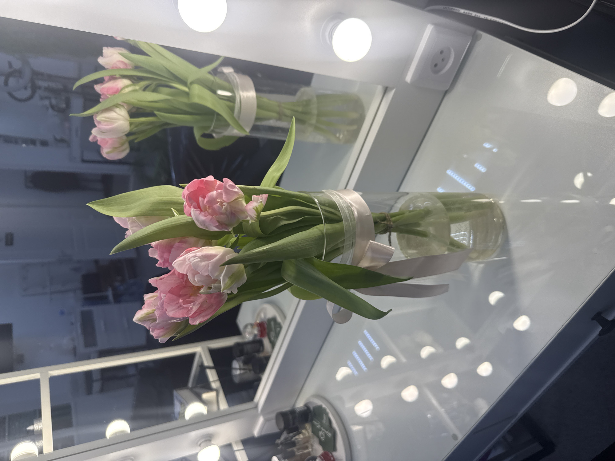 A still-life photo shows a vase of pink and white tulips placed on a vanity table with a built-in mirror framed by light bulbs. The vanity is located in Chaikovsky, Russia.
The tulips are arranged in a clear glass vase tied with a pale pink ribbon. The flower arrangement includes multiple blooms and green leaves. The vase appears to be partially filled with water.
The vanity has a glossy white surface. The mirror is outlined with numerous white light bulbs. Reflections of the flowers and other objects are visible in the mirror. An electrical outlet can be seen in the upper right corner of the mirror. Some cosmetic items are visible on the counter below the vanity. The scene is well-lit, suggesting a studio or indoor setting.