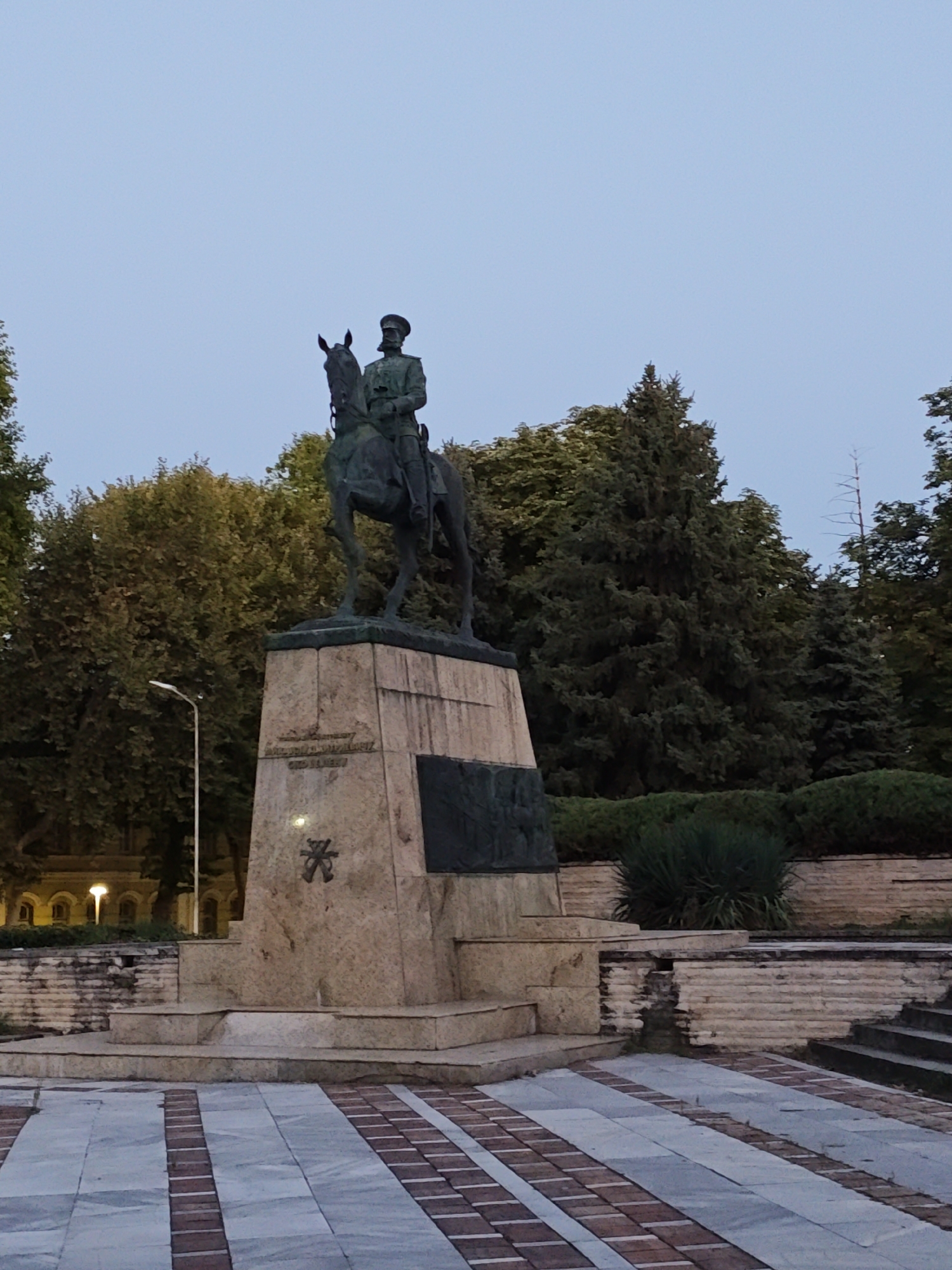 The image shows an equestrian statue in the city of Pleven, Bulgaria. The statue depicts a man in military uniform on horseback, mounted on a large stone pedestal. The pedestal has inscriptions and decorative elements. The statue is located in an outdoor setting, with trees and bushes visible in the background. The sky is overcast, suggesting it is either early morning or late evening. The foreground features a paved area with a patterned design of light and dark grey tiles with interspersed red brick lines. There are also stone steps and retaining walls in the scene, indicating a public space or park. A lamppost is visible to the left of the statue.