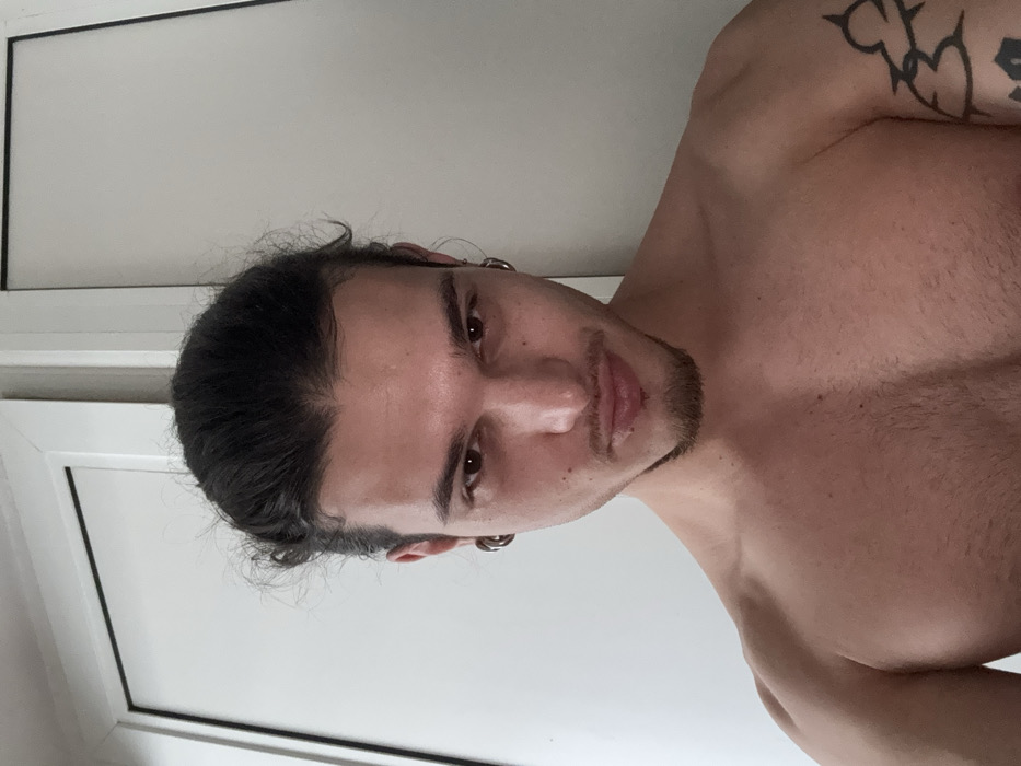 An indoor, close-up image features a single bare-chested male individual looking directly into the camera. The subject has dark hair tied back in a bun or top knot, a goatee, and dark eyes. He wears black tunnel earrings in both earlobes and two small silver hoop earrings in the upper cartilage of his right ear. An abstract black tattoo is visible on his upper left arm. His shoulders and upper chest show muscle definition. The background consists of plain white vertical surfaces, appearing to be two adjacent doors or a wall with a door frame. The lighting is even. No specific landmarks or architectural features are present to identify a precise location within the city of Sofia, Bulgaria; the setting is a generic interior space.