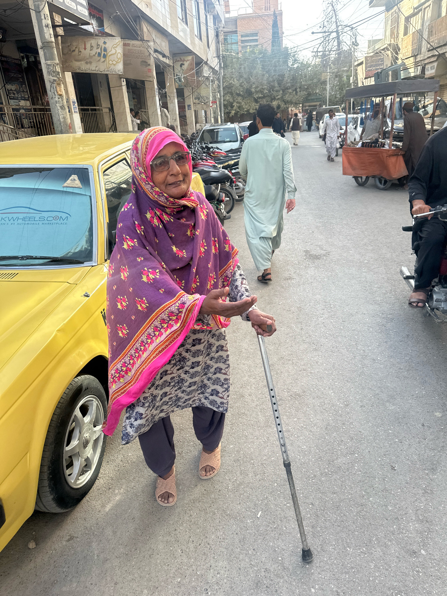 A woman wearing glasses and a pink and purple headscarf is standing on a street in Quetta City Tehsil, Pakistan, holding a walking stick. She is wearing a grey and white patterned top and dark blue pants. To her left is a yellow car. In the background, other people are walking and riding motorcycles, and there are shops and buildings lining the street. The scene appears to be during the daytime.