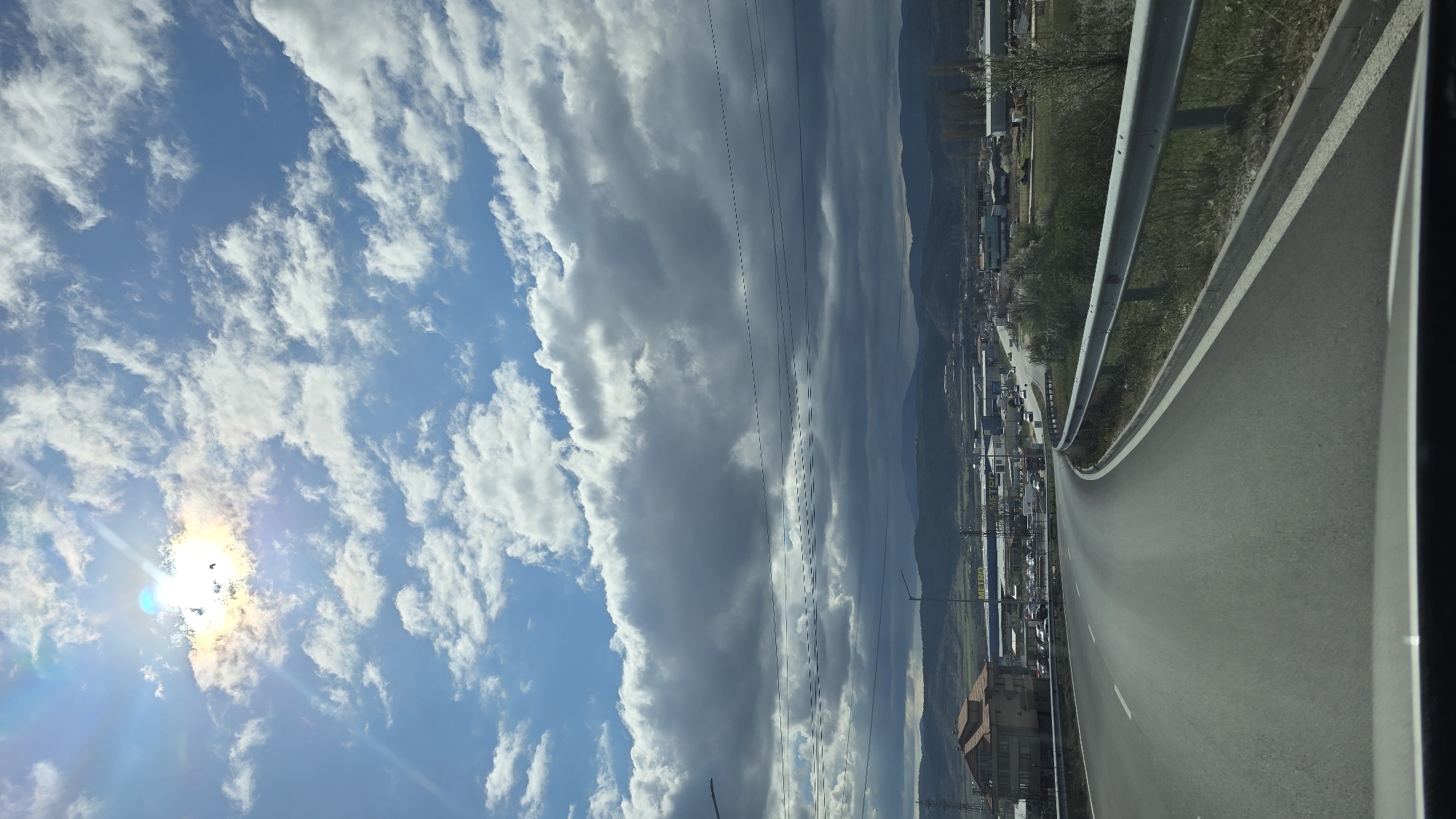 The image depicts an asphalt road with a metal guardrail, presumably a highway or major roadway, curving to the right of the frame. Buildings and vegetation are visible alongside the road, suggesting an urban or suburban setting. In the background, a mountain range is partially obscured by clouds. The sky above is a mix of blue and white, with a bright sun on the left. Power lines are strung across the view. The image is oriented sideways, with the sky on the left and the road on the right. The location is Blagoevgrad, Bulgaria.