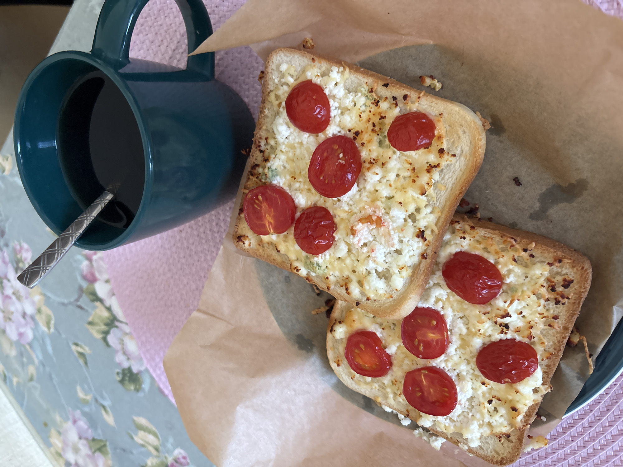 Two pieces of toasted bread are topped with a white cheese mixture and sliced cherry tomatoes. The toast is placed on brown parchment paper, which sits on a pink textured placemat. To the right of the toast is a dark teal mug filled with black liquid, presumably coffee, with a silver spoon inside. The background shows a floral tablecloth. The image is a still life of food and a beverage, likely taken indoors in a domestic setting, no specific location in Naberezhnye Chelny, Russia can be identified from the image.