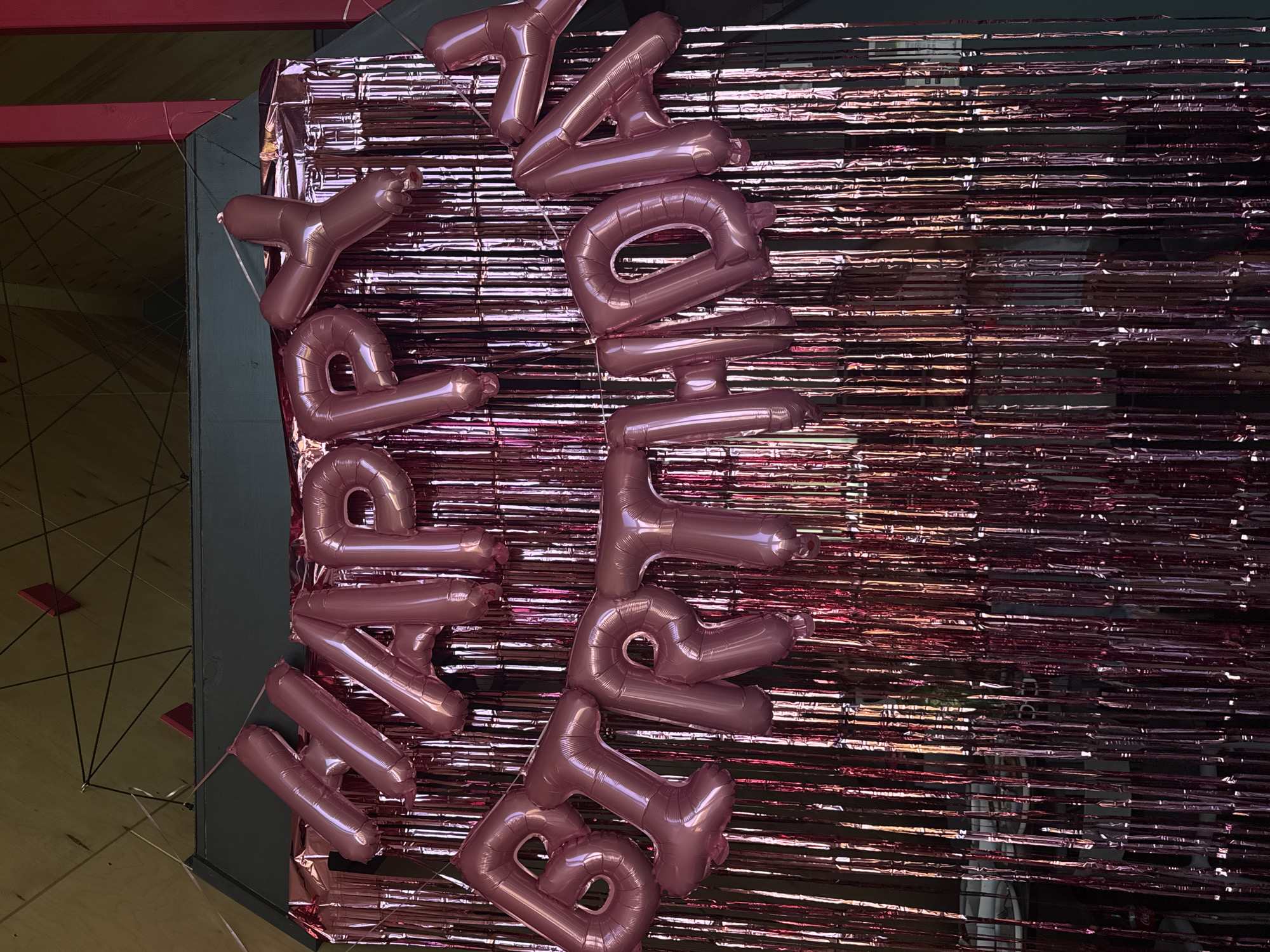 The image shows pink "Happy Birthday" balloons arranged vertically against a shiny pink tinsel backdrop. The background is partly obscured. The location is not specified.