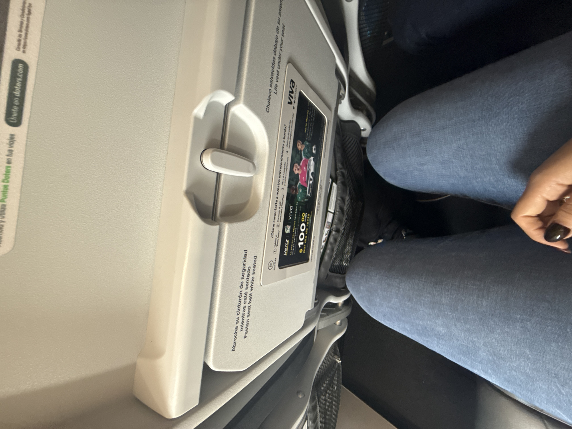 The image shows an airplane seat with a tray table in the upright position. The tray table has instructions and advertisements, including a Hertz advertisement offering car rentals for $100 per day. Part of a passenger wearing blue jeans is also visible. The image does not contain information to place it in San Antonio, United States of America.