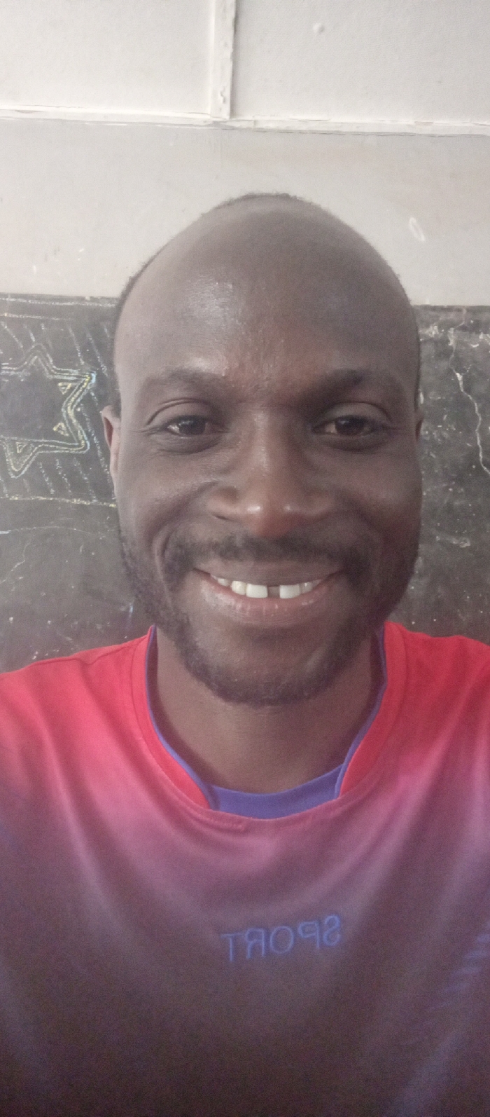 An adult male subject is depicted in a close-up, head-and-shoulders portrait, positioned indoors in Barnawa, Nigeria. The individual has a dark complexion, a receding hairline, and maintains a short beard and mustache. He is smiling directly at the camera, displaying a visible diastema between his two upper front incisors. He wears a short-sleeved garment, primarily reddish-pink/coral, with a blue/purple contrasting collar visible underneath. The word "SPORT" is legible in reverse text on the lower front portion of his shirt. The background to the left of the subject features a dark surface with a white chalk outline of a Star of David. To the right, the background consists of a lighter, possibly textured wall. The upper background shows a white ceiling or upper wall section with visible structural lines.
