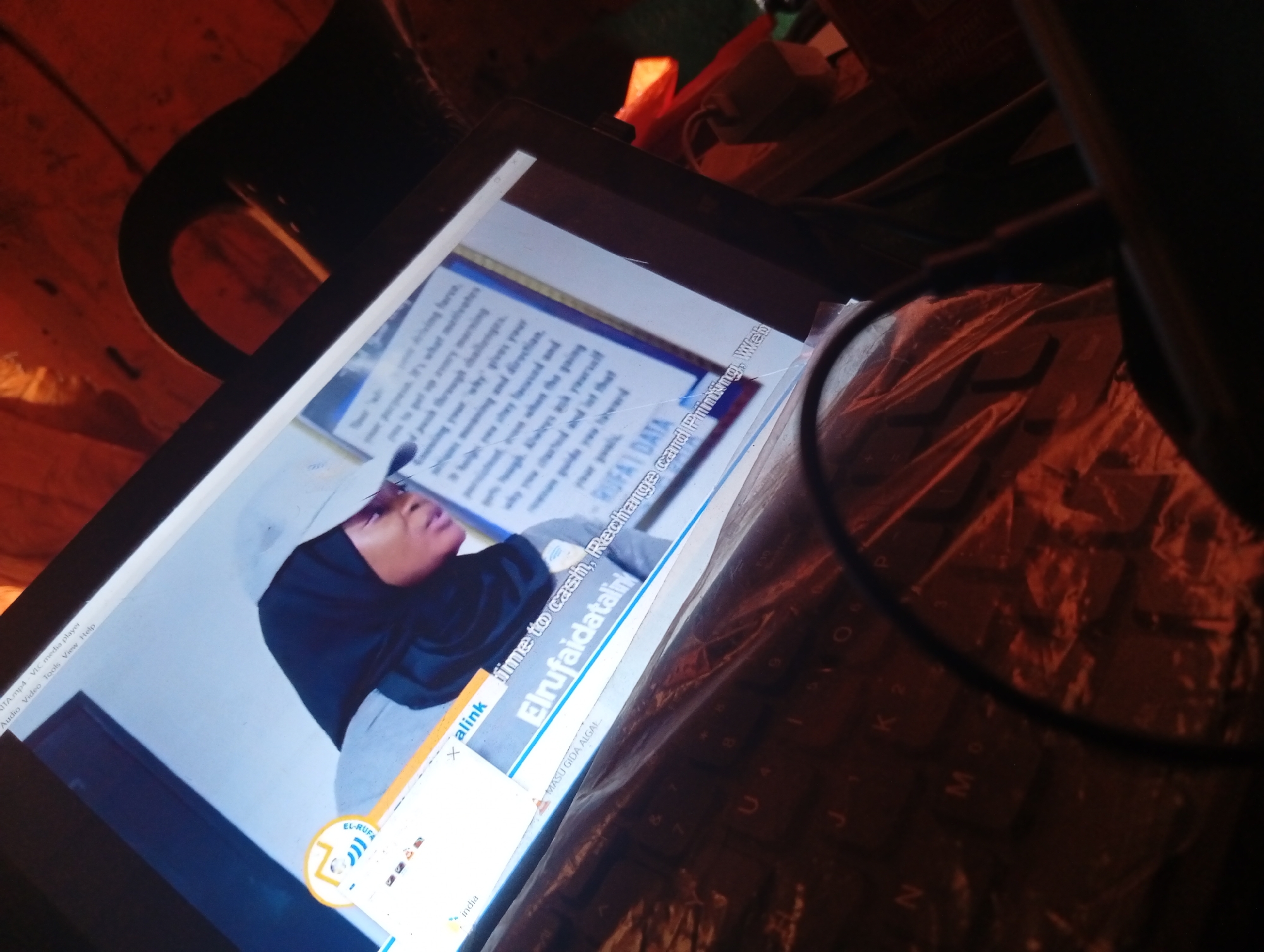 The image shows a tablet computer displaying a video with a person wearing a headscarf. The screen content includes text in English and Arabic, with a website address visible. The tablet is positioned on a keyboard, with a dark, circular object, possibly a lens or a part of a microphone, obscuring a portion of the scene. The background is dimly lit and appears to be an indoor environment, with some objects that suggest a workspace or a home setting. The overall lighting is warm and orange-toned. There are no visible indications of the city of Zongoma Kyari, Nigeria.