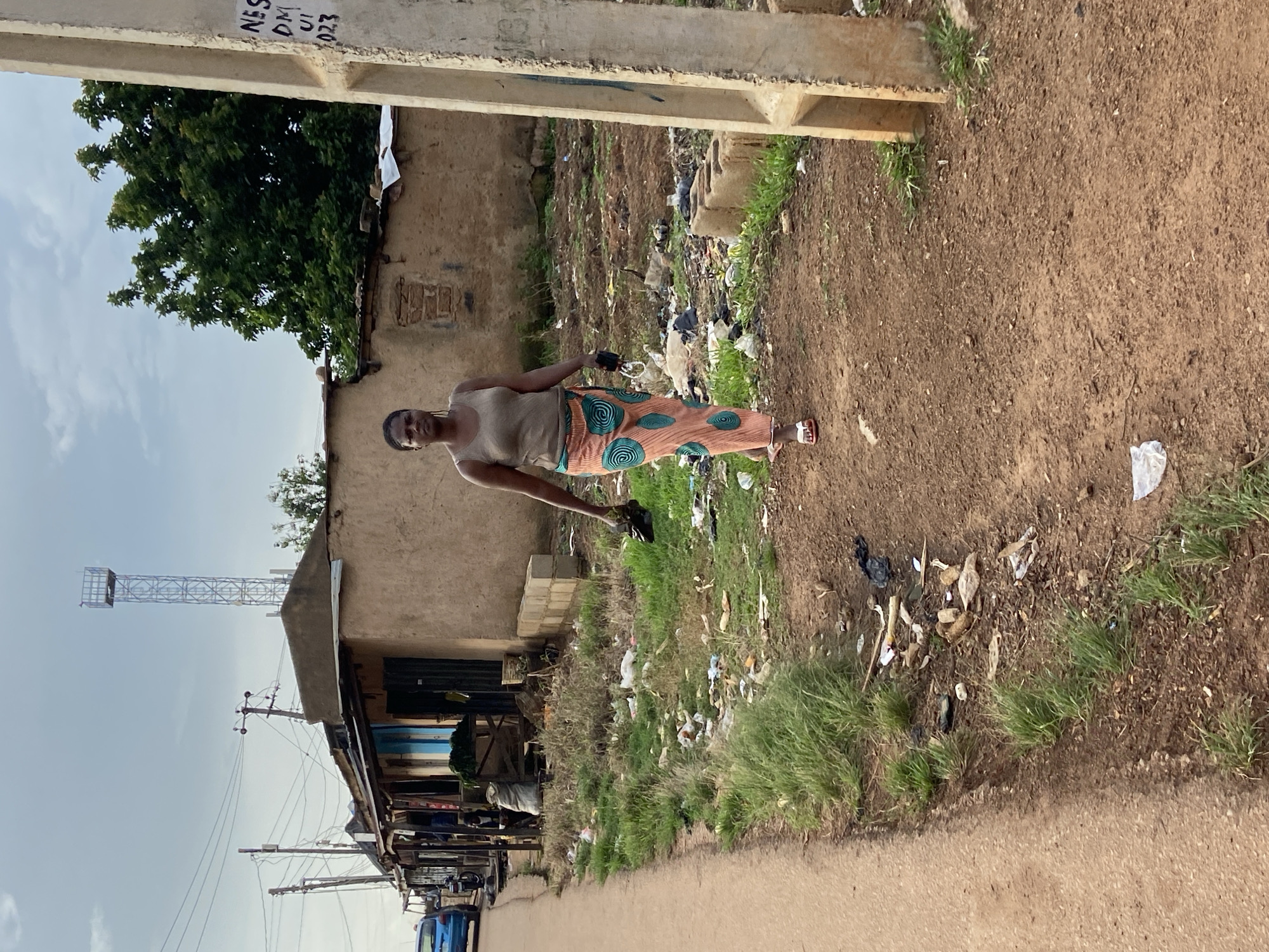 In Du, Nigeria, a woman walks on a dirt path beside a roadside that is littered with trash. The woman is wearing a light-colored tank top and a long, patterned skirt. She is carrying a black bag in her right hand and a pair of black sandals in her left hand. She is walking away from the camera, towards a small cluster of buildings that appear to be shops or homes. The buildings are constructed from a mixture of mud and concrete. Behind the buildings, a tall cell tower can be seen, and trees with dense foliage are visible. The sky is overcast, suggesting cloudy weather. The roadside also features grass and more trash. A blue car is parked on the left side of the road.
