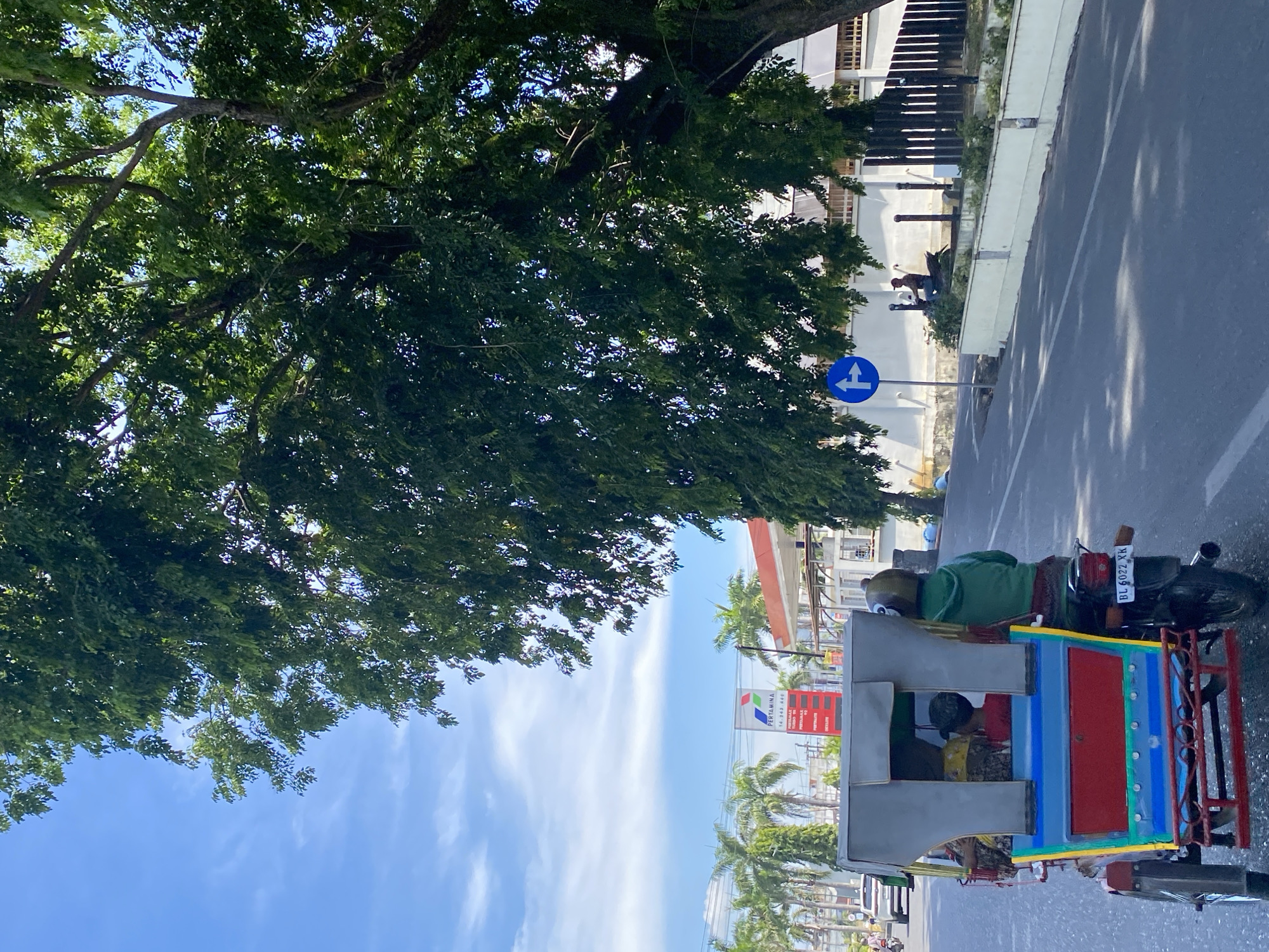 An eye-level, medium shot captures a colorful, three-wheeled passenger vehicle, known as a "becak," traveling on a paved road under a bright, partly cloudy sky. The becak, painted blue, red, and yellow, is driven by a person in a green hooded jacket, who wears a helmet. Three passengers are visible in the back of the becak. On the left side of the road, a large tree with dense green foliage dominates the upper right portion of the frame, casting shadows on the road. In the background, palm trees line the left side of the road, and a gas station with a prominent "Pertamina" sign is visible. A blue traffic sign indicating a mandatory right or straight turn is mounted on a pole. Further down the road, a person on a motorcycle is stopped near a white wall and fence. The scene appears to be in Lhokseumawe, Indonesia, based on the presence of the Pertamina gas station and the style of the becak.