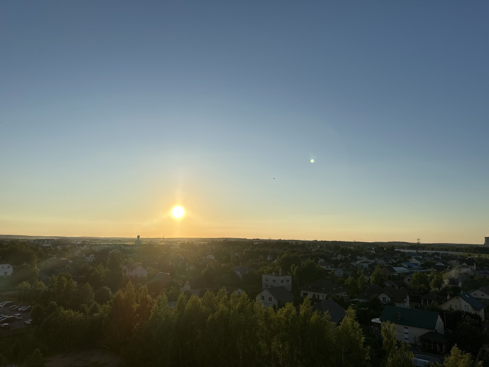 A panoramic view of a suburban or semi-rural area is captured during sunset, likely in or near the Шчомысліцкі сельскі Савет, Belarus. The sky transitions from a deep blue at the top to a soft yellow-orange glow near the horizon, indicating the setting sun. The sun itself is a bright, luminous orb positioned in the left half of the image, casting a warm light across the landscape. A fainter, smaller circular light, possibly a lens flare or a reflection, is visible in the upper right quadrant of the sky.
Below the sky, a dense canopy of green trees dominates the middle ground, interspersed with houses and buildings. The architecture appears to be predominantly residential, with pitched roofs and varying colors, suggesting a mix of individual homes and possibly some smaller apartment buildings. The trees are lush and full, creating a natural barrier between the sky and the residential areas.
In the background, a more distant cityscape or industrial area is discernible, with taller structures and a more developed skyline. A prominent water tower is visible on the left side of the distant horizon. The overall impression is one of a peaceful evening scene in a developed yet green area. There are no visible people or animals in the frame. The image is taken from an elevated perspective, looking down on the scene.