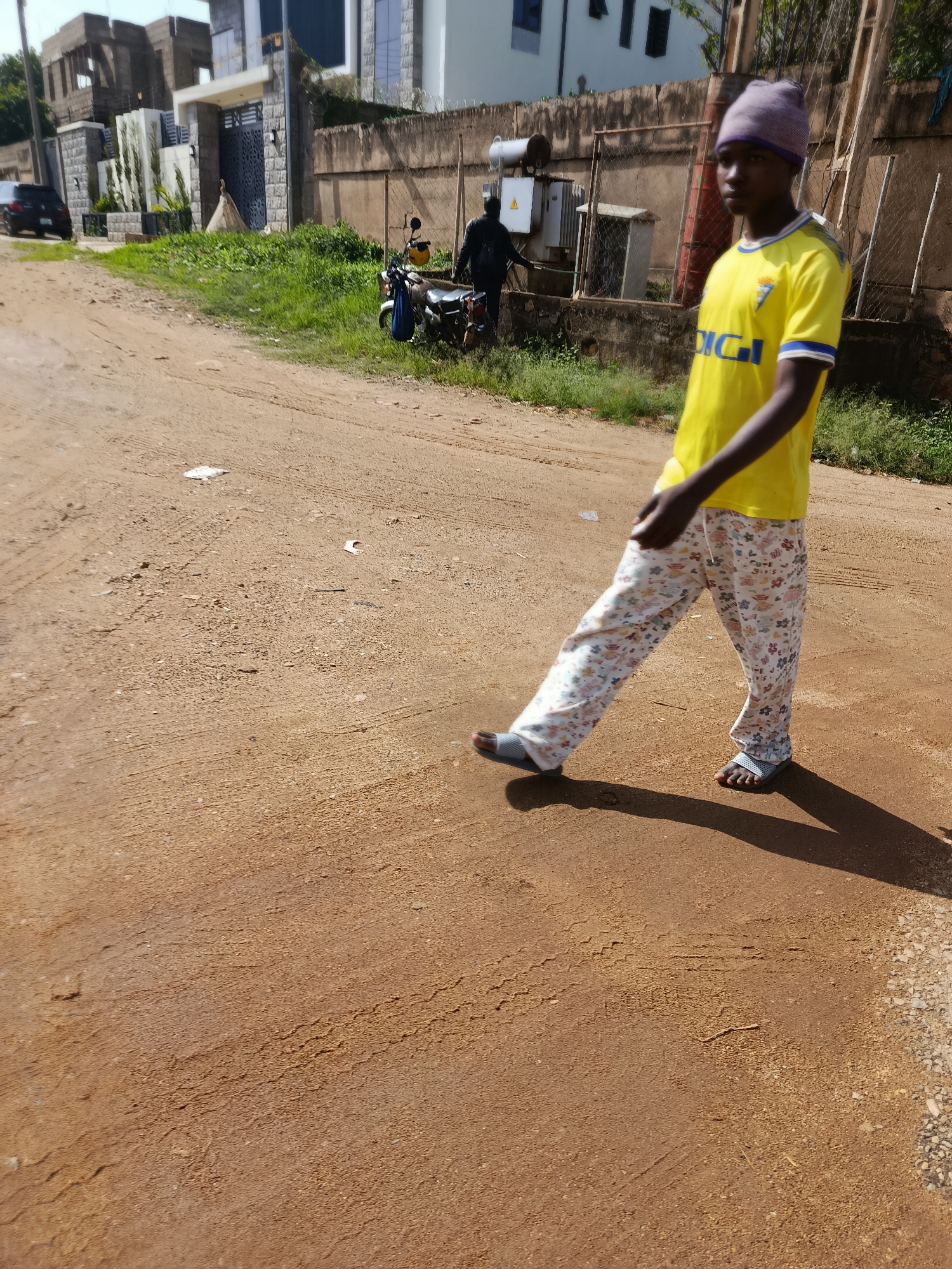 A young boy walks on a dirt road in Rafin Par, Nigeria. He is wearing a yellow t-shirt with blue trim, patterned pajama pants, and grey sandals. In the background, there are buildings, a parked car, and a motorcycle. The boy is walking towards the right of the frame, casting a shadow on the road. The environment appears to be an unpaved street in a residential area.