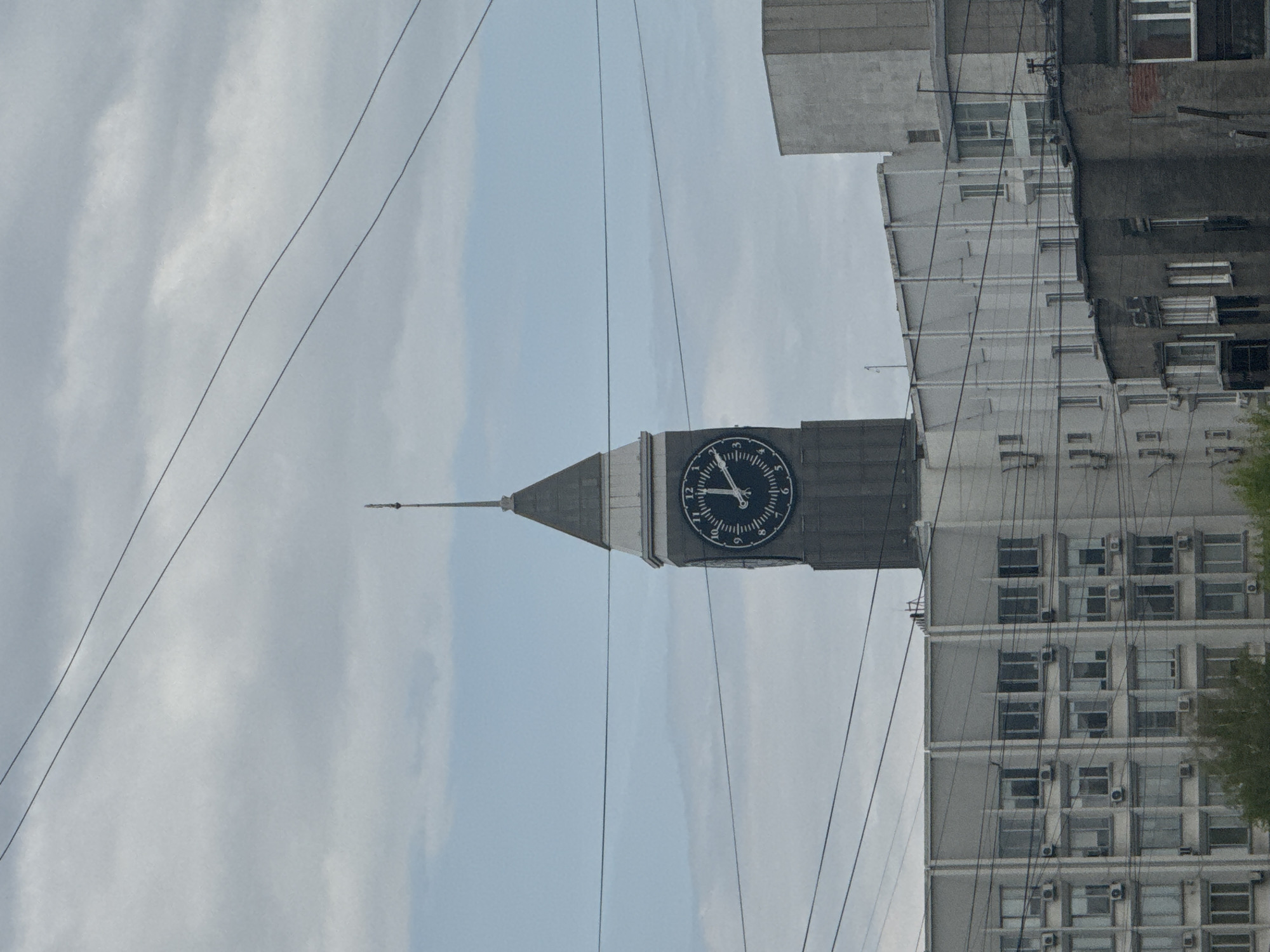 The image depicts the Krasnoyarsk Clock Tower in Russia against a cloudy sky. The tower is positioned vertically, featuring a prominent clock face on its rectangular structure, topped with a pointed spire. Adjacent to the tower is a multi-story building, characterized by rows of windows and air conditioning units. The scene includes numerous power lines intersecting the view between the tower and the sky. No people are visible.