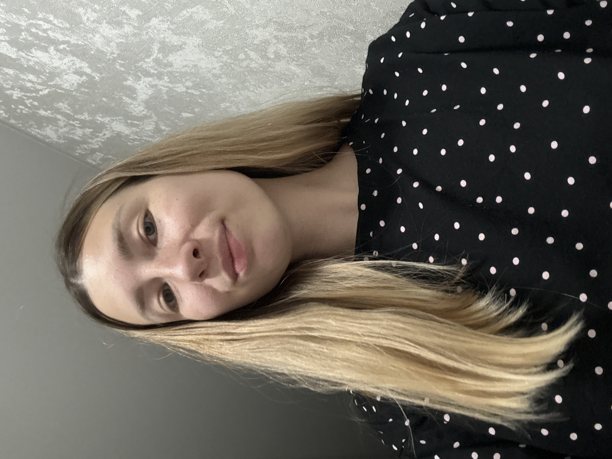 A woman with long, blonde hair is wearing a black, polka-dotted shirt. The background consists of a textured, light-colored wall in the upper portion and a solid, gray wall in the lower portion. There is no indication of Moscow, Russia, or a city in general, as the scene appears to be indoors.