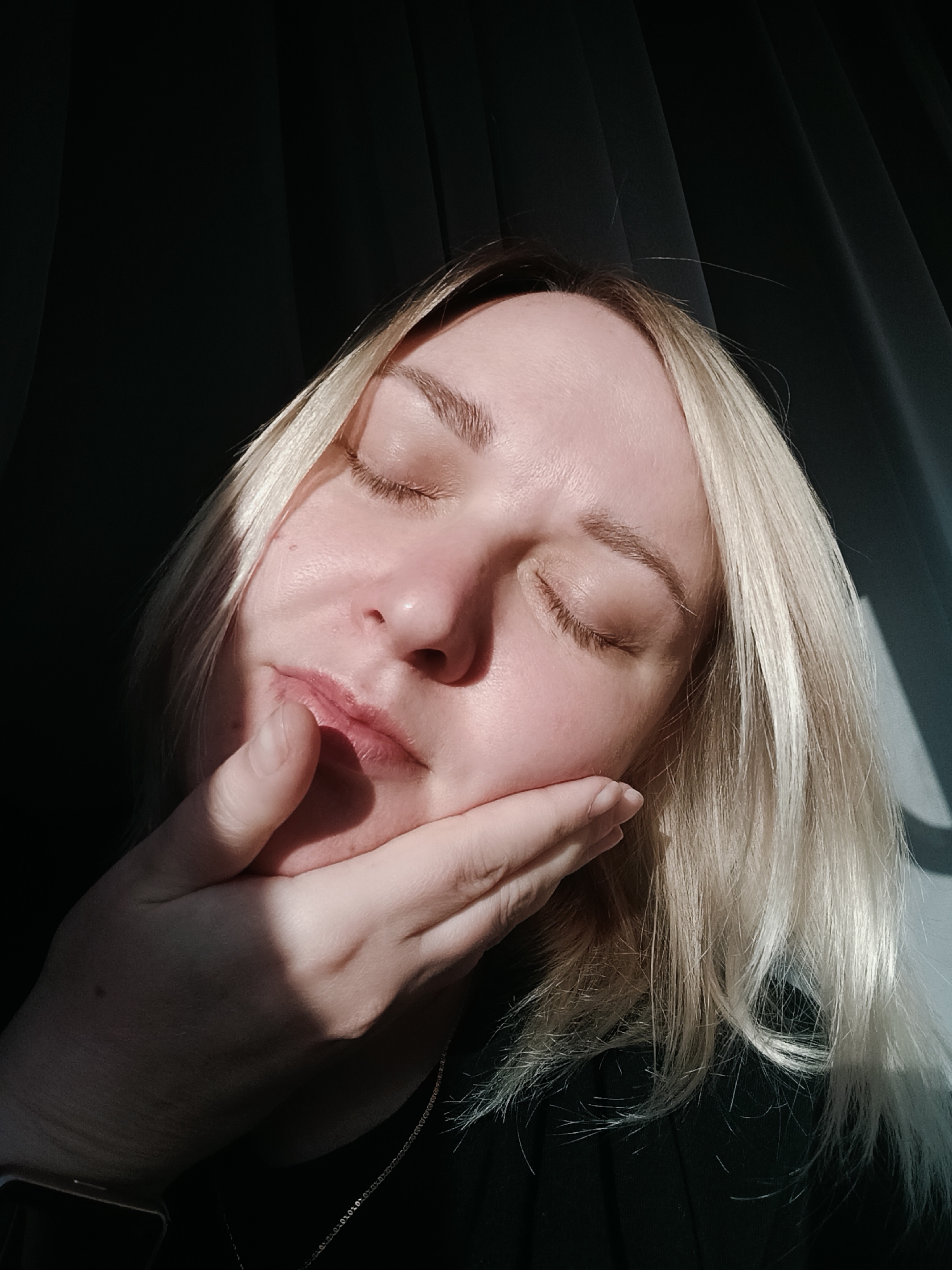 A close-up, sunlit portrait captures a fair-skinned woman with blonde hair in a peaceful, contemplative pose. Her eyes are closed, and her head is tilted slightly as she rests her chin on her left hand. Direct, strong sunlight illuminates the right side of her face and hair, creating a stark contrast with the deep shadows falling across the left side of her face, the underside of her hand, and the dark, draped background, likely curtains.
The woman's blonde hair is highlighted by the light, showing variations in tone and texture. Her skin, visible in the direct light, shows natural texture and a few minor blemishes. She wears a dark top, and a delicate silver chain necklace is visible around her neck. On her left wrist, partially obscured by shadow, a smartwatch can be seen. The background consists of dark, vertically draped fabric, emphasizing the subject and the dramatic lighting. The overall impression is one of quiet repose or deep thought in a domestic setting, likely during the daytime when sunlight is streaming indoors. No visible text is present.