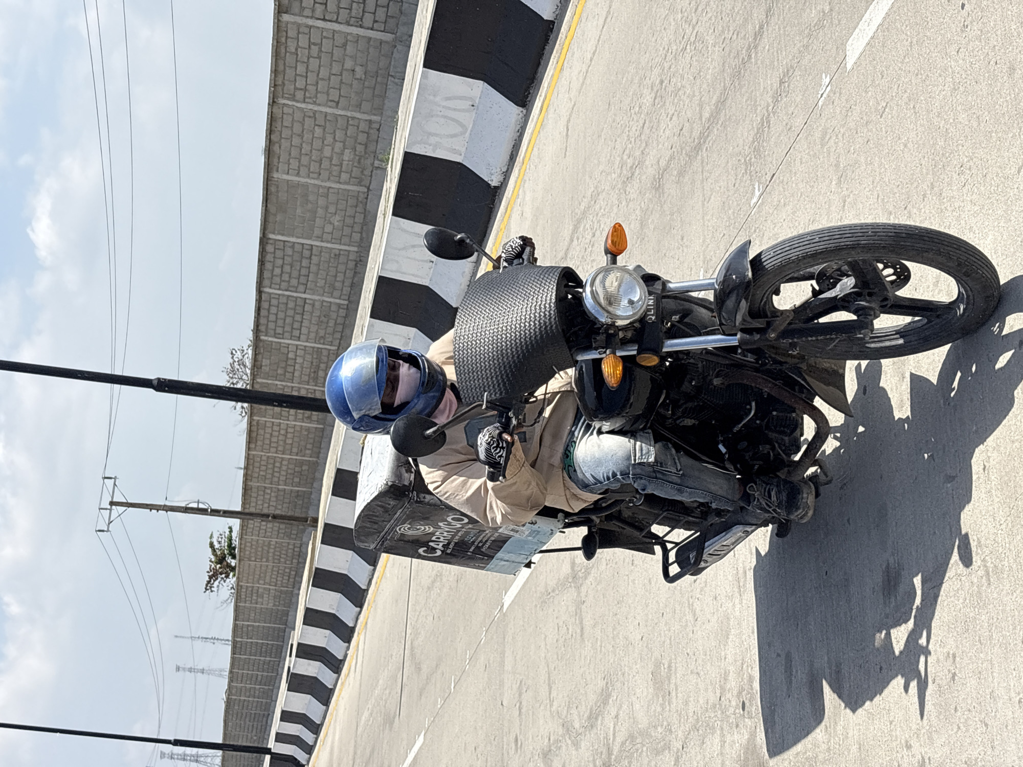 A man in a blue helmet rides a black motorcycle on a gray concrete road. The motorcycle has a basket on the front and a delivery bag labeled "CARRIGO" on the back. He wears a light brown jacket and jeans. A black and white striped barrier is to the man's left. Power lines and utility poles are in the background. The image appears to be taken in Igboefon, Nigeria.