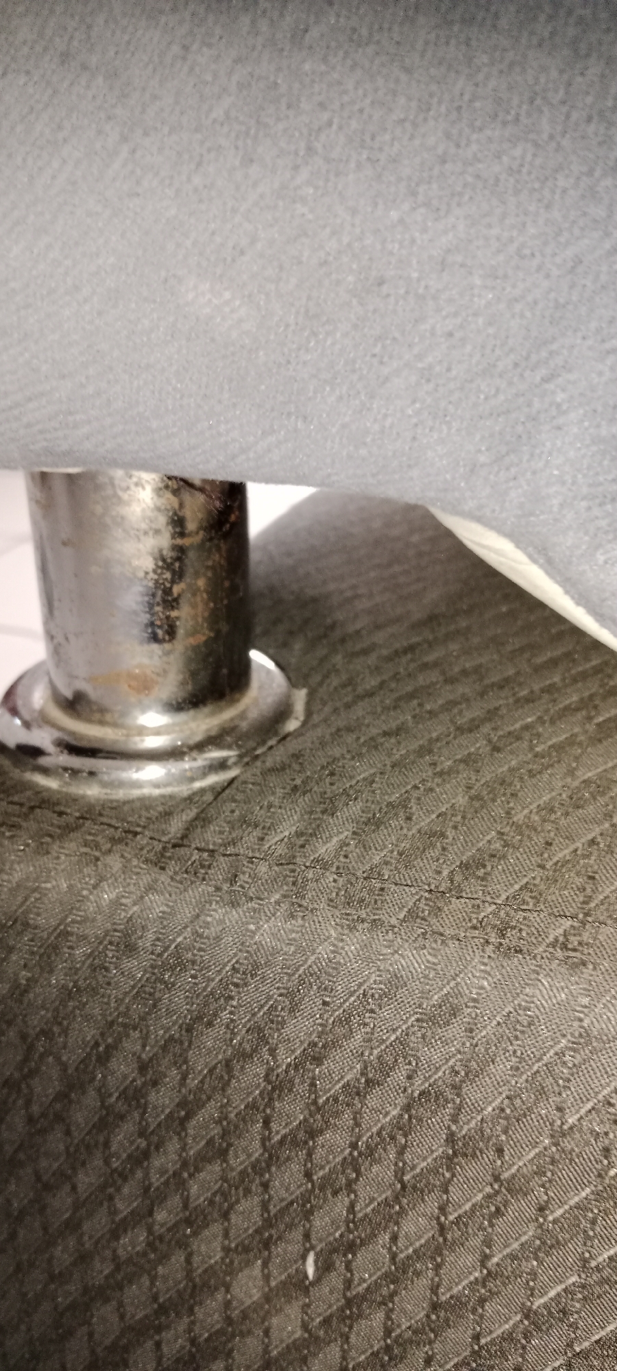 The image shows a close-up view of a metal support leg, likely from a piece of furniture, resting on a textured dark fabric. The metal leg is cylindrical and appears to have some rust or discoloration on its surface. It has a flared base that sits on the fabric. The fabric has a repeating diamond-shaped pattern and is dark gray or black. Above the metal leg, a lighter gray fabric is visible, possibly another part of the furniture. The scene appears to be indoors, and the lighting is dim. There are no people or animals visible in the image. There is no information in the image to suggest the location is Subang, Indonesia.