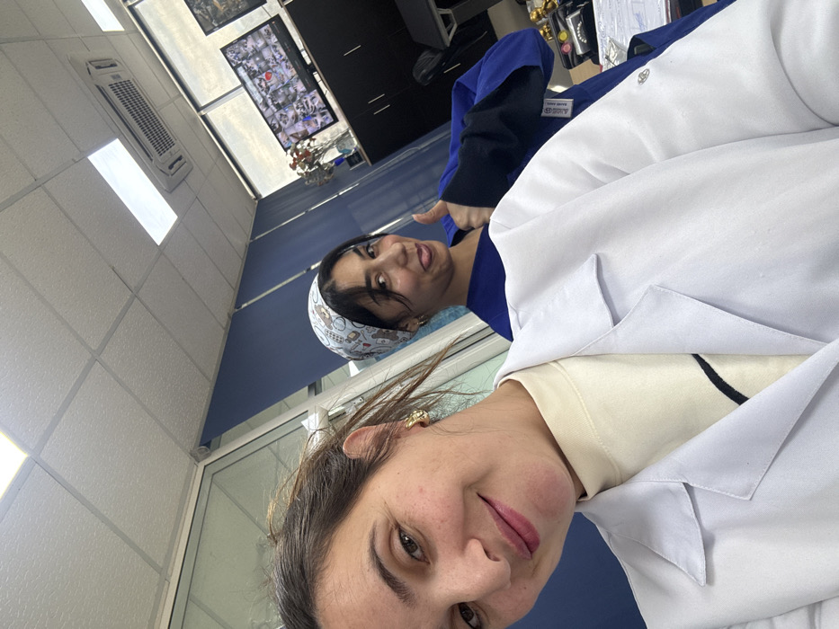 The image depicts two women inside an indoor professional or medical facility located in Meknes, Morocco. The woman in the foreground, whose upper torso and head are visible, has light-to-medium skin, dark hair pulled back, and is wearing a white lab coat over a cream-colored turtleneck shirt. She has gold hoop earrings and is smiling subtly towards the camera. Behind her, a second woman, visible from the chest up, has darker hair and skin. She is wearing a light blue scrub cap with white patterns and a blue scrub top, with black long sleeves visible underneath. This woman is captured making a playful face, sticking her tongue out, and giving a thumbs-up gesture with her right hand.
The environment includes a white drop ceiling with recessed fluorescent lighting and an overhead air conditioning unit. A glass partition is visible on the left side of the frame. In the background, a dark-colored credenza or cabinet supports a flat-screen monitor displaying a grid of multiple surveillance camera feeds. A vase containing red flowers is positioned on the credenza next to the monitor. Partially drawn blue window blinds are visible in the background, suggesting exterior windows. The precise location within Meknes is not identifiable from visual cues in the image.
