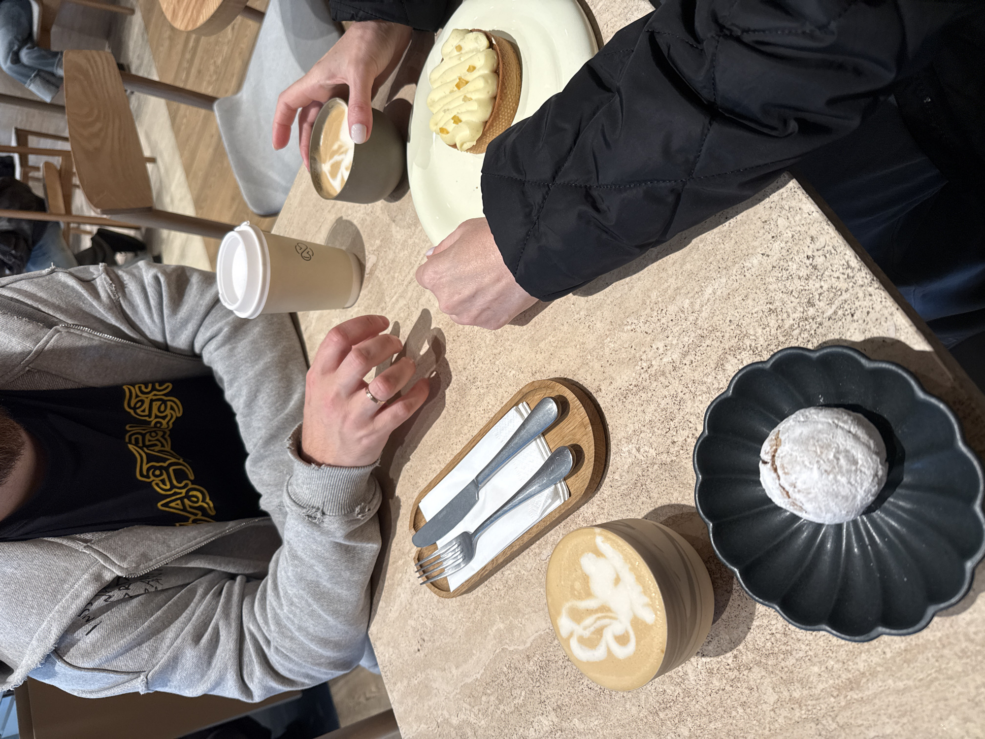 The image shows an overhead view of a table with two people. One person is wearing a gray hoodie over a black t-shirt and a ring on their finger. The other person is wearing a black puffer jacket. On the table are two cups of coffee, a pastry, a plate with a fork and knife, and a powdered donut on a black plate. Based on the cafe decor and style of food, it is possible that this image was taken at a coffee shop in Moscow, Russia.