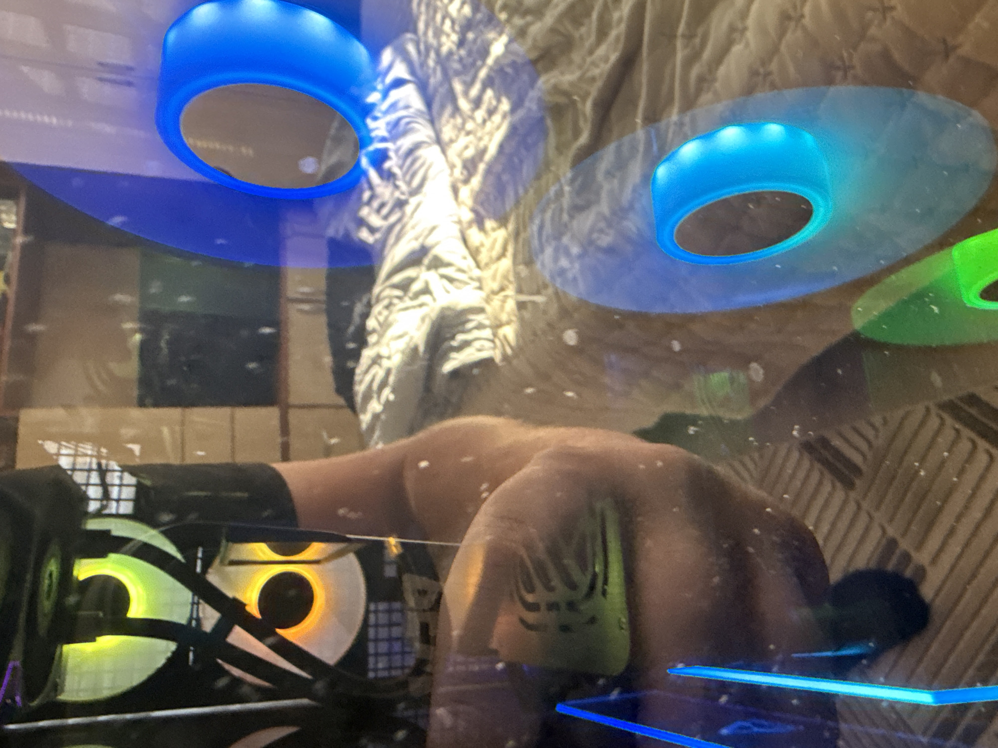 The image shows a close-up view of a person's arm and torso reflected in a glass surface. The reflection reveals several computer fans with RGB lighting, displaying green and orange hues. On the right side of the image, a bed with a patterned quilt is visible in the background, partially obscured by the reflection of the fans and some blue, ring-shaped lights. The environment appears to be indoors, possibly a room with a computer setup. There is no indication of the location being in Draginovo, Bulgaria.