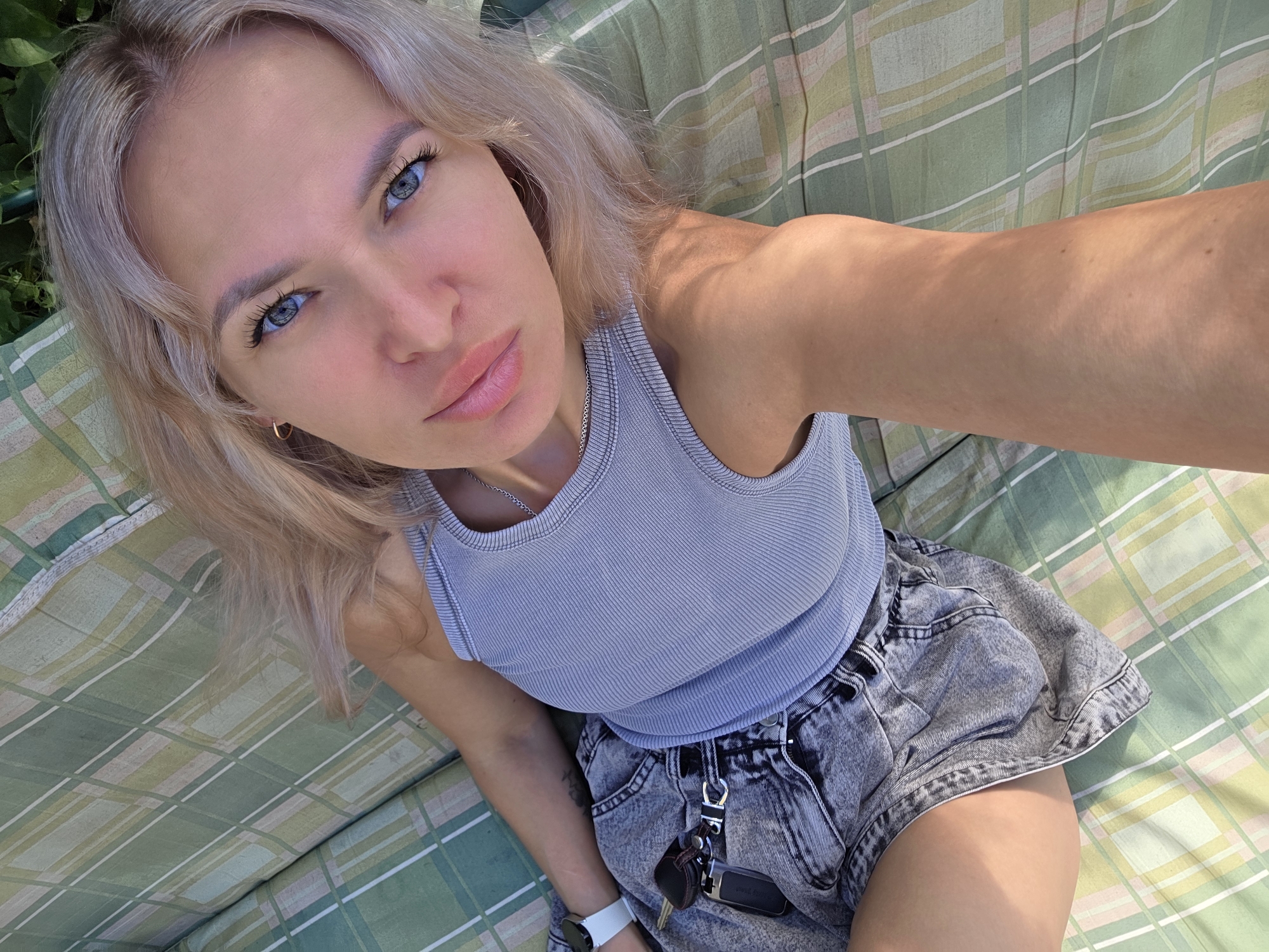 The image shows a young woman with blonde hair and blue eyes lying down on a green and white plaid outdoor cushion. She is wearing a grey ribbed tank top and denim shorts. A watch is visible on her left wrist, and car keys with a brown leather fob are attached to her shorts. The background consists of greenery and what appears to be foliage, suggesting an outdoor setting. There is no specific indication of the city of Chaikovsky, Russia, within the image.