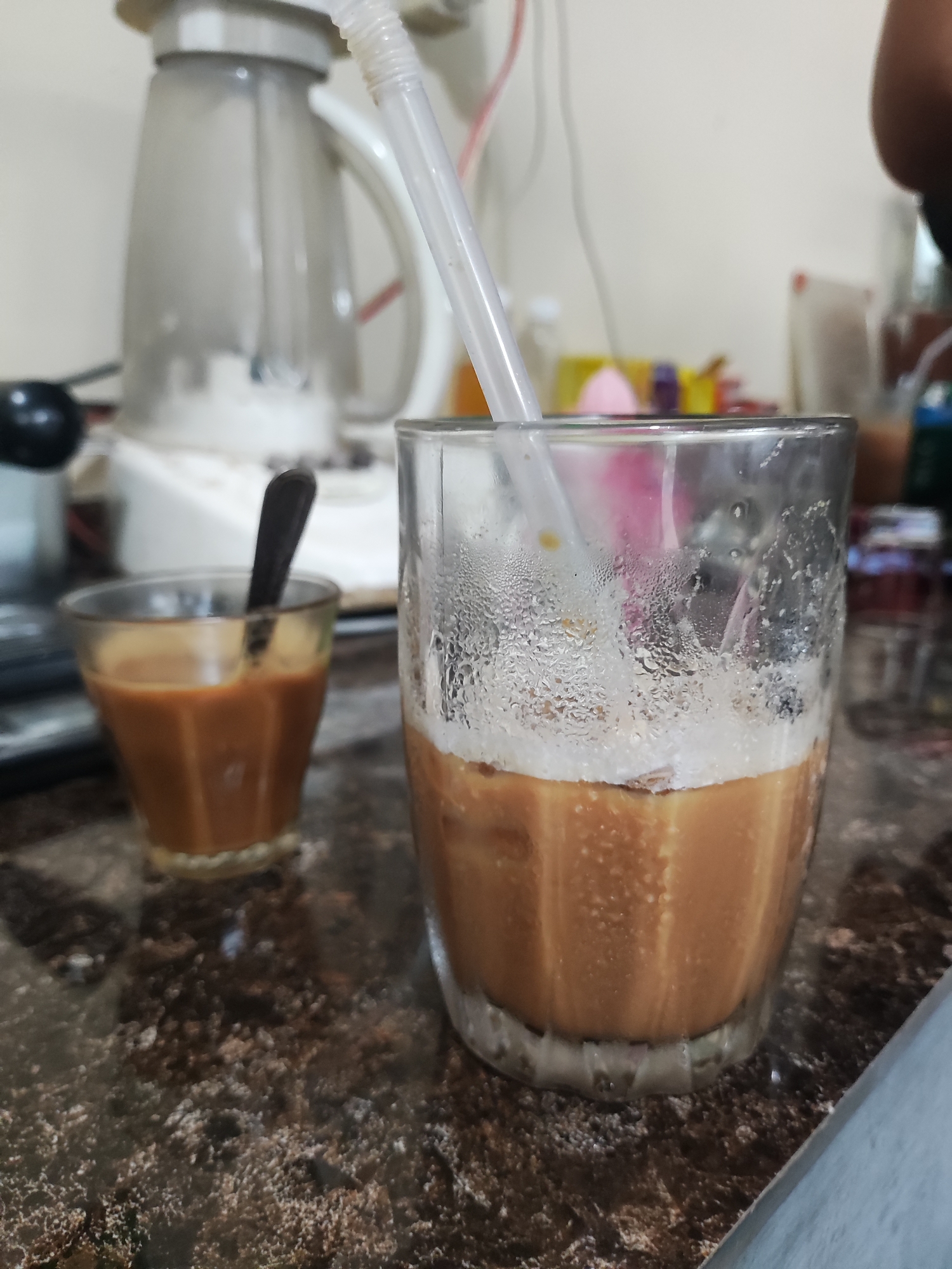 The scene depicts two glasses of milky coffee on a dark brown countertop. The glass on the right has a straw and condensation on its outer surface. The smaller glass on the left has a spoon in it. Behind the glasses, there is a blender and assorted items, including what appears to be packaging. The overall setting suggests a kitchen or a cafe, but the exact location within Lhokseumawe, Indonesia, cannot be determined based solely on this image. There is no direct evidence of specific people or interactions.