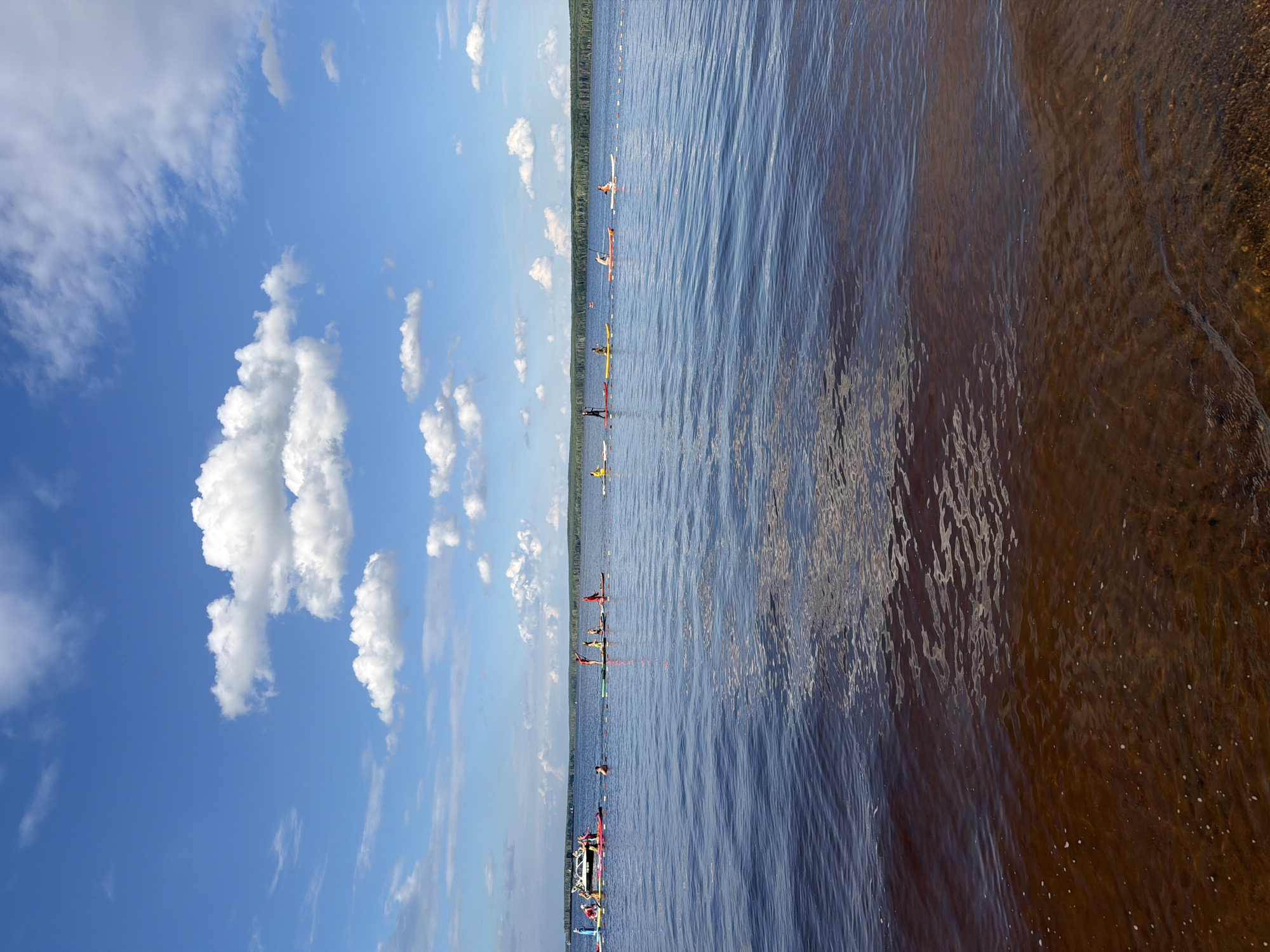 The image depicts a group of people in kayaks on a wide, calm body of water, likely a lake or river. The scene is set outdoors under a bright blue sky dotted with fluffy white clouds, suggesting fair weather and daytime. In the foreground, the shallow water near the shore reveals a sandy bottom with some small pebbles. The water surface is rippled, reflecting the sky and creating dynamic patterns.
Further out on the water, several kayakers are spread out, appearing to be participating in some form of organized activity or race. They are in various positions on the water, with some closer to the shore and others further out. The kayaks are of different colors, including white, yellow, and red. Some of the kayakers are wearing brightly colored clothing.
In the distance, a dense line of trees forms the horizon, indicating a wooded shoreline. The overall atmosphere appears to be relaxed and active, with people enjoying water sports on a pleasant day. No visible text is present in the image. The location context suggests this could be a recreational area or a venue for water-based events in Chaikovsky, Russia.