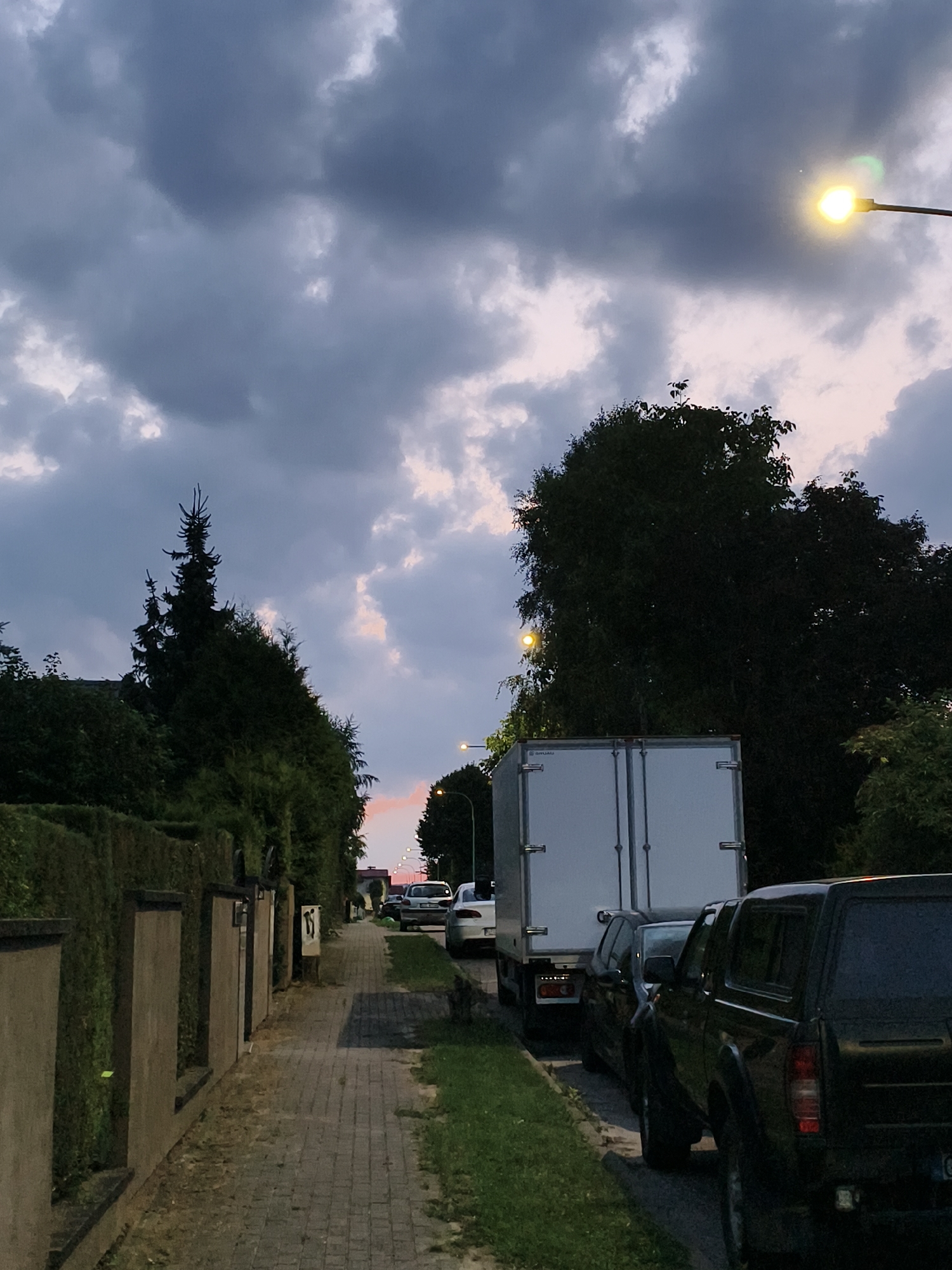 The image depicts a street scene in Tczew, Poland, at dusk. A sidewalk made of interlocking pavers runs along the left side of the image, bordered by a low wall and neatly trimmed hedges. Several parked vehicles are visible on the street: a dark-colored pickup truck with a cap, a dark sedan, and a white truck trailer. Further down the street, other cars and a lamppost are discernible. The sky is filled with dramatic, dark clouds, with a hint of pink and orange from the setting sun visible near the horizon. A street lamp on the right side of the image is illuminated. The overall impression is of a quiet residential or suburban street as evening approaches.