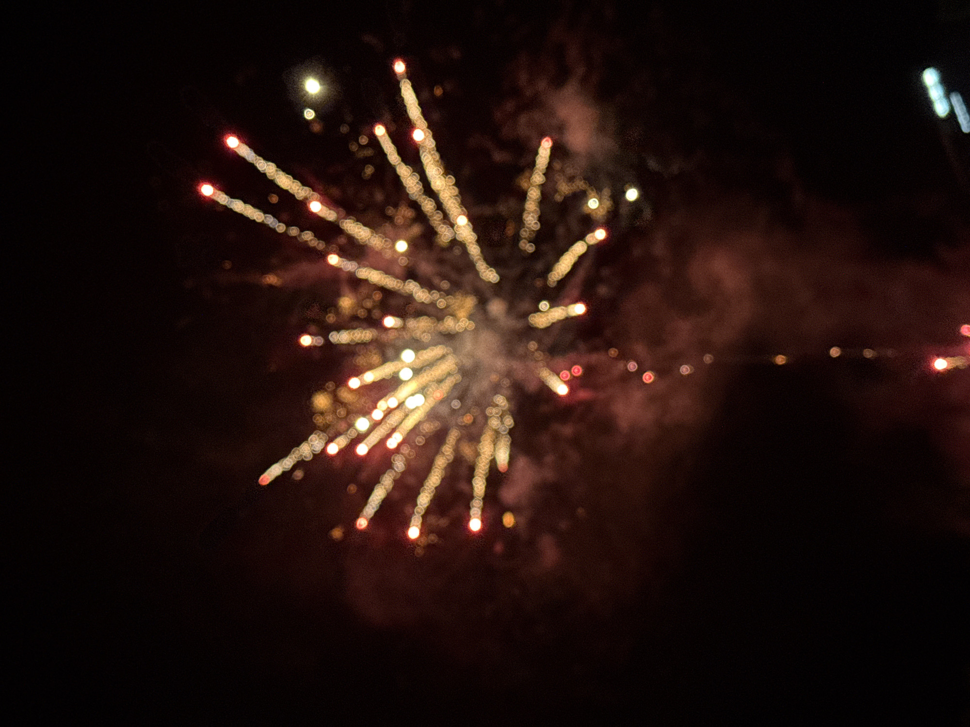 This forensic image captures a nocturnal scene in Debrashtitsa, Bulgaria, depicting a fireworks display against a dark sky. The central focus is a luminous burst of golden-yellow and red light trails, extending radially outward, characteristic of an exploding pyrotechnic device. The light sources exhibit a distinct bokeh effect, appearing as blurred circular or oval shapes due to the shallow depth of field. Reddish-brown smoke and haze are visible within the dark background, particularly around the core of the fireworks burst and trailing downwards. A singular, faint, glowing stream extends from the lower portion of the primary explosion towards the bottom of the frame. The environment is uniformly dark, consistent with nighttime conditions, with no discernible people, structures, or specific landscape features. A few extremely blurred, distant artificial lights are faintly visible in the far background, primarily in the upper right quadrant.
