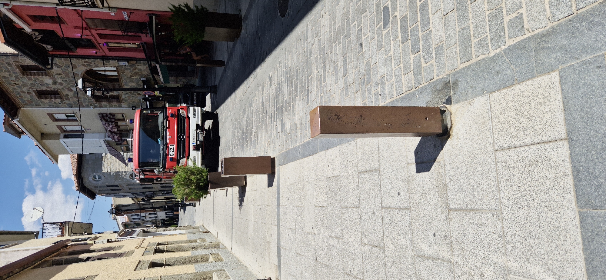 The image shows a street scene in Navaluenga, Spain. A red fire truck is parked on a cobblestone street lined with buildings. There are planters with green bushes along the sidewalk, and bollards are installed to prevent vehicles from entering certain areas. The sky is blue with some clouds. The buildings appear to be made of stone and stucco, with some having balconies. The scene is well-lit by sunlight, casting shadows on the ground. There are no people clearly visible in the image.