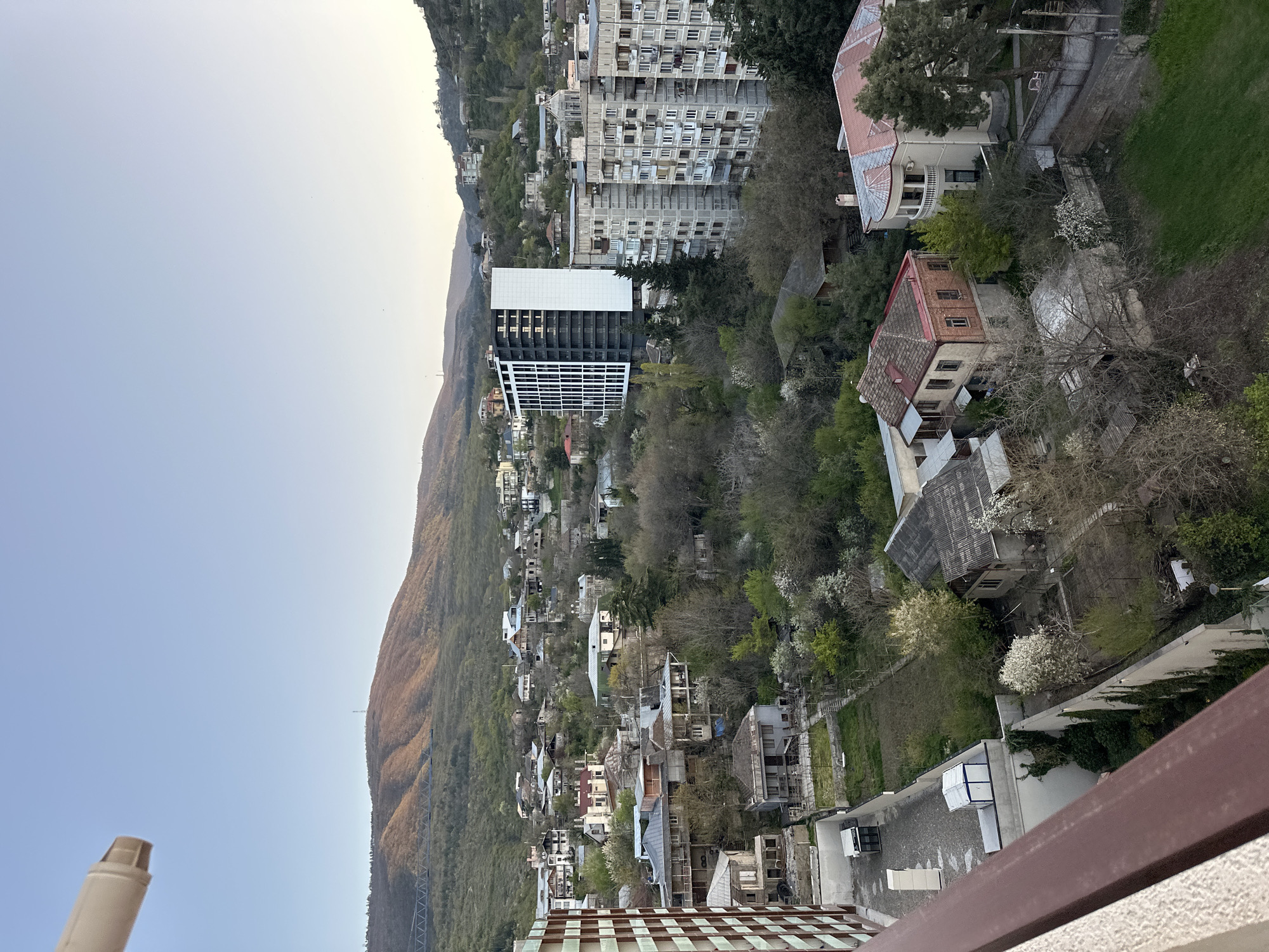 The image shows a hillside neighborhood in Tbilisi, Georgia. The houses are interspersed with trees and vegetation. A mountain rises in the background. Several tall buildings are visible in the distance. The sky is light blue. A balcony railing is in the foreground.