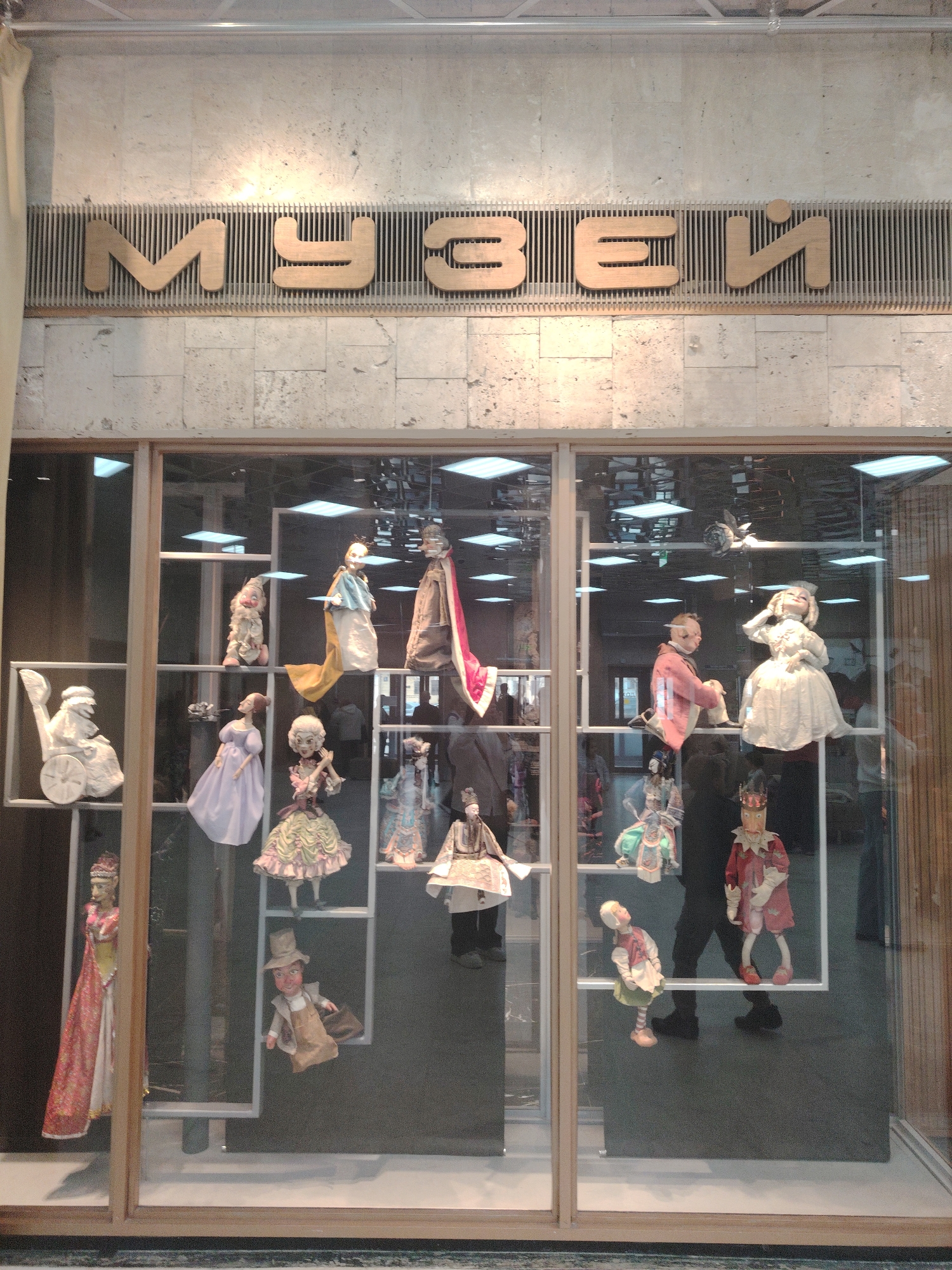 The image captures a museum display case, possibly located in an unknown city. The case contains a collection of vintage dolls and puppets dressed in various historical and theatrical costumes. Above the display, the word "МУЗЕЙ" (Museum) is written in large, stylized gold letters, set against a textured concrete wall with a ventilation grill. The interior of the museum is visible through the clear glass of the display case, reflecting a series of overhead lights and the silhouettes of people walking by, including a person in dark attire who seems to be observing the display. The scene is well-lit, with a mix of natural and artificial light creating reflections on the glass surfaces.