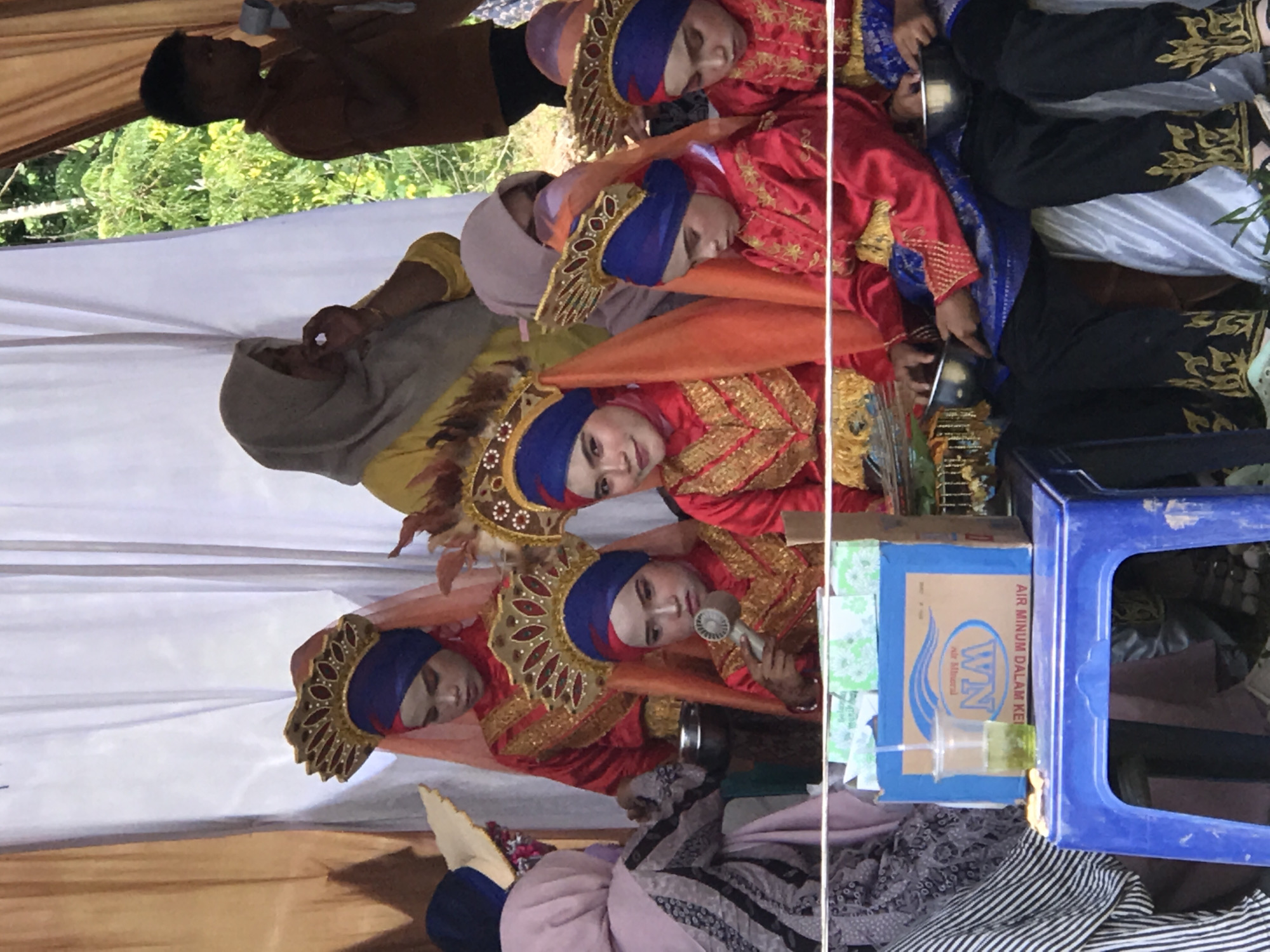 In Langkuta, Indonesia, a group of young people are dressed in traditional attire, likely for a performance or cultural event. The scene appears to be outdoors, with a backdrop of white curtains and trees visible in the distance. Several individuals, particularly young women, are adorned in vibrant red and gold costumes, complete with ornate headpieces and veils. They are seated on a low blue plastic stool. In the foreground, a blue cardboard box with the "WIN AIR MINUM DALAM KEMASAN" logo and a glass of green liquid with a straw are placed on the stool. One individual is holding a small, metallic bowl. Another person, a young boy in a brown shirt, is visible in the background, holding a white object. The overall atmosphere suggests a festive or ceremonial occasion.
