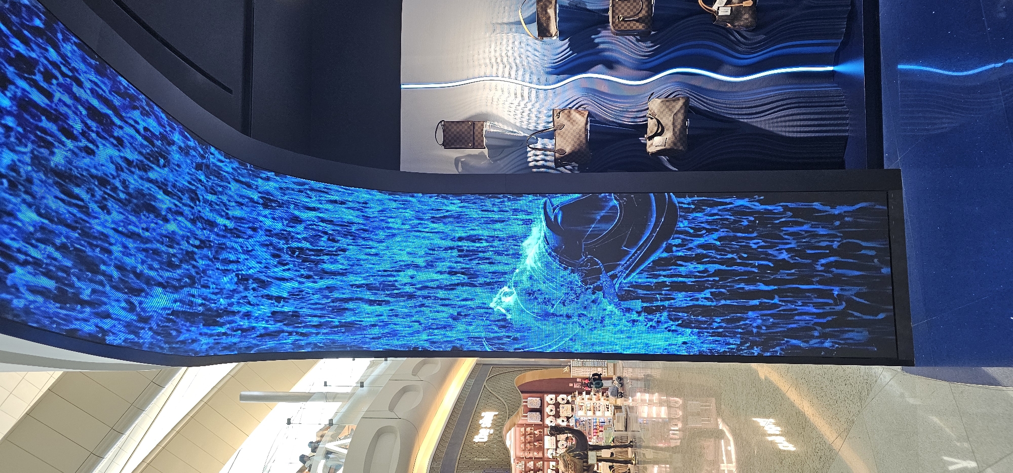 The image depicts a curved LED screen displaying a dynamic blue digital waterfall or abstract pattern, possibly at Al Matar airport in the United Arab Emirates. To the right of the screen, a retail display features several handbags, seemingly from a luxury brand, arranged on shelves against a dark, undulating background illuminated by blue light. The handbags appear to be made of brown checkered material. In the background, the spacious interior of a modern building is visible, with architectural elements like elevated walkways, railings, and lighting. A large retail store with a gold-trimmed facade and various products displayed behind glass is also present. A camel statue and some shoppers can be observed in the distance, further indicating a public space like an airport or a large shopping mall. The overall scene suggests a commercial and high-traffic environment.