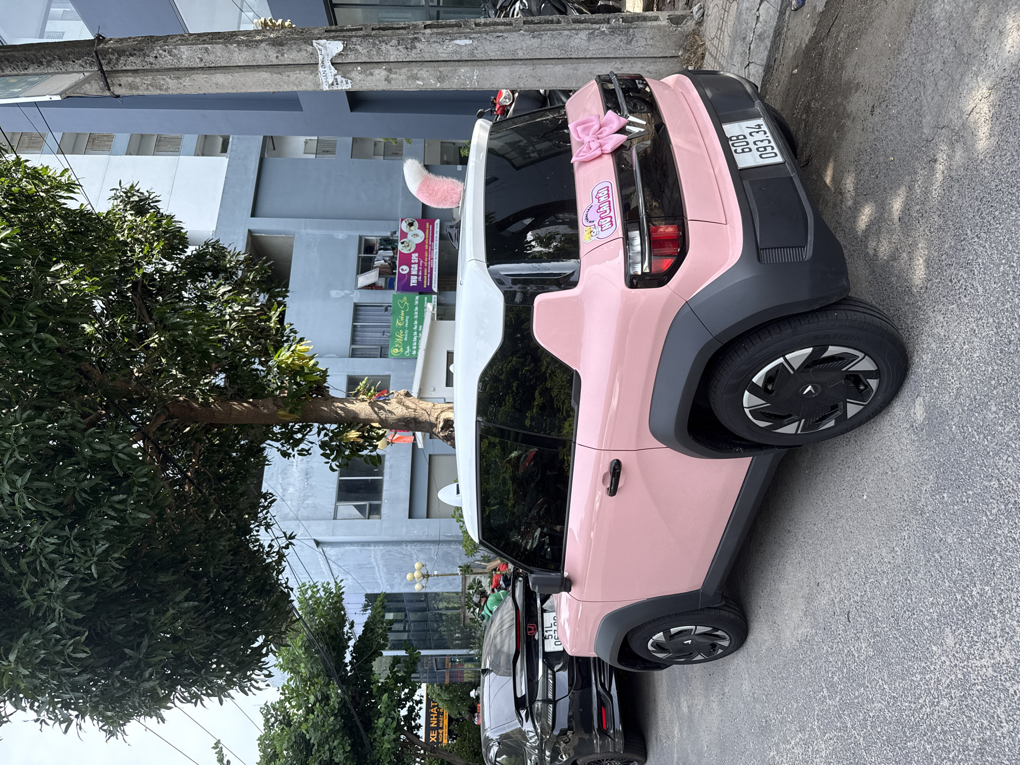 A pink electric vehicle with a white roof is parked on a city street in Nam Nha Trang Ward, Vietnam. The car has a pink bow and a fluffy white and pink tail attached to its rear, as well as what appear to be bunny ears on its roof. A black car is parked behind it. Buildings line the street, and a utility pole stands to the right of the vehicles. Trees provide shade over the scene.