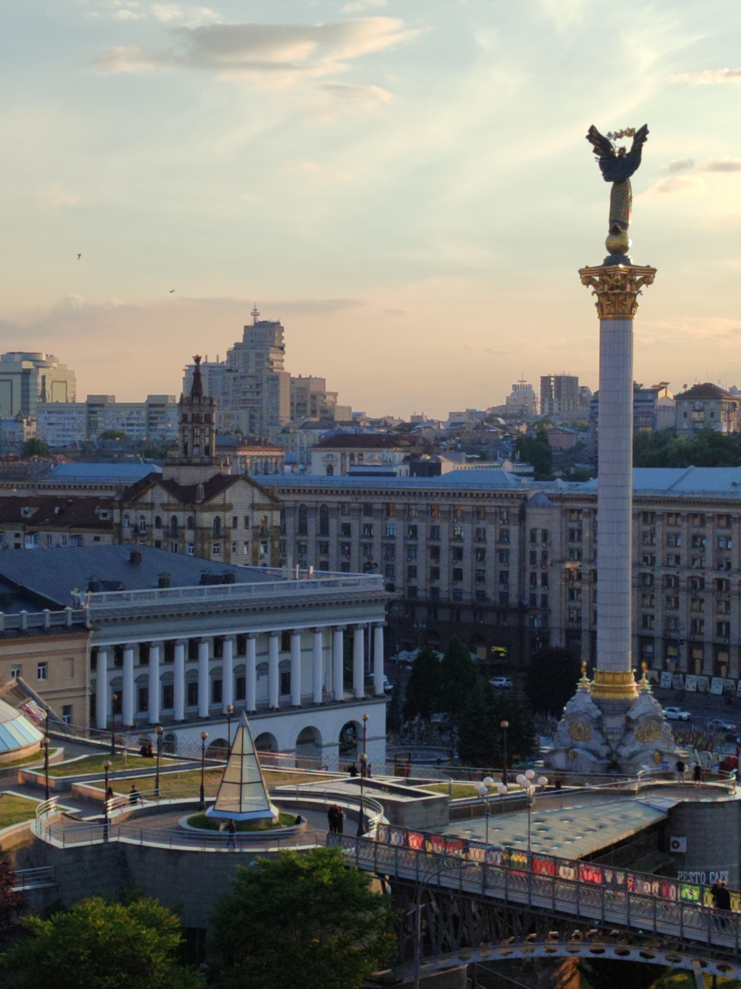 This image captures a vibrant cityscape, likely at golden hour, either late afternoon or early morning, indicated by the soft, warm light bathing the scene and the gentle pink, orange, and pale blue hues in the sky. Scattered clouds drift overhead, and two small birds are visible in flight.
The focal point is the towering white Independence Monument (Monuments of Independence of Ukraine) on the right, adorned with a golden base and capital, topped by a bronze statue of Berehynia, a female figure with outstretched wings holding a wreath. This monument strongly identifies the location as Maidan Nezalezhnosti (Independence Square) in Kyiv, Ukraine.
The surrounding area reveals a blend of architectural styles. In the mid-ground left, a grand building with numerous white classical columns stands prominently. Behind it and stretching into the background are various multi-story buildings, some appearing older and ornate, others more functional high-rises. In the immediate foreground, an elevated plaza features a striking glass pyramid structure, which serves as an entrance to an underground mall. A modern pedestrian bridge, adorned with what appear to be colorful banners or flags, spans across the lower right, connecting different levels of the square.
A few people are sparsely visible, walking on the pedestrian bridge and in the plaza around the glass pyramid, engaged in everyday activities. The overall atmosphere is peaceful and serene, showcasing a bustling city settling into twilight or awakening to a new day. A partially visible sign on the bridge railing reads "PESTO CAFE."