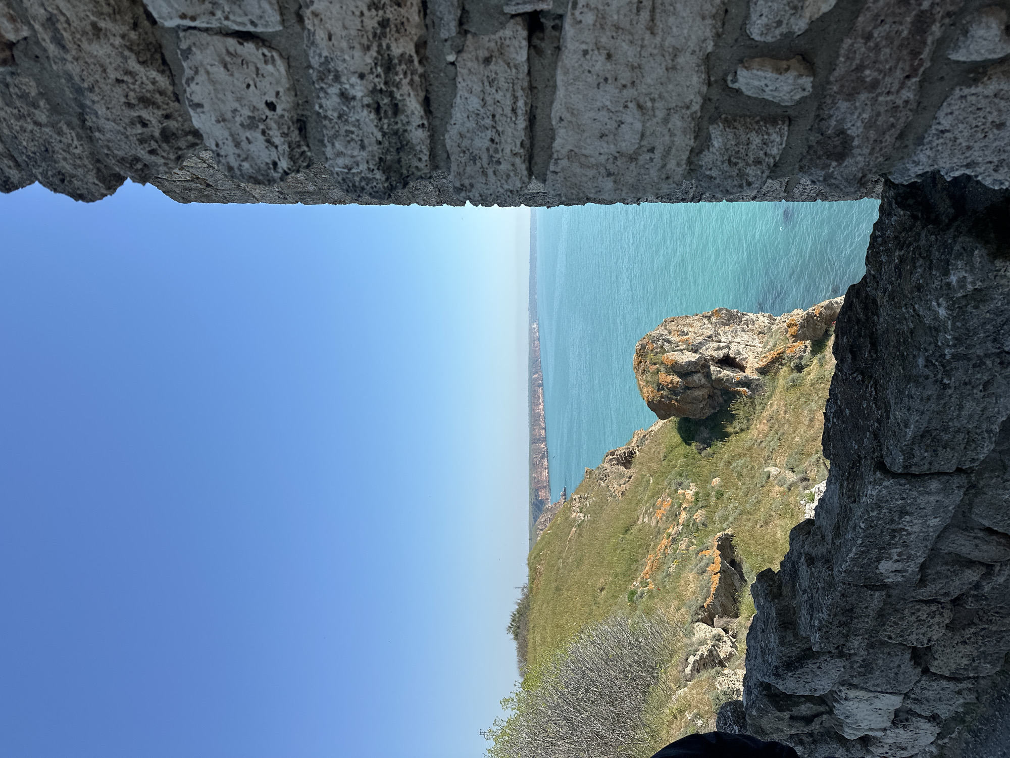 The image shows a view from a stone structure in Balgarevo, Bulgaria. The frame is formed by stone walls at the top and right side of the image. The view looks out over the Black Sea, with a coastline visible in the distance. A rocky cliffside covered with grass and vegetation is seen on the right, sloping towards the sea. The sky is clear blue.