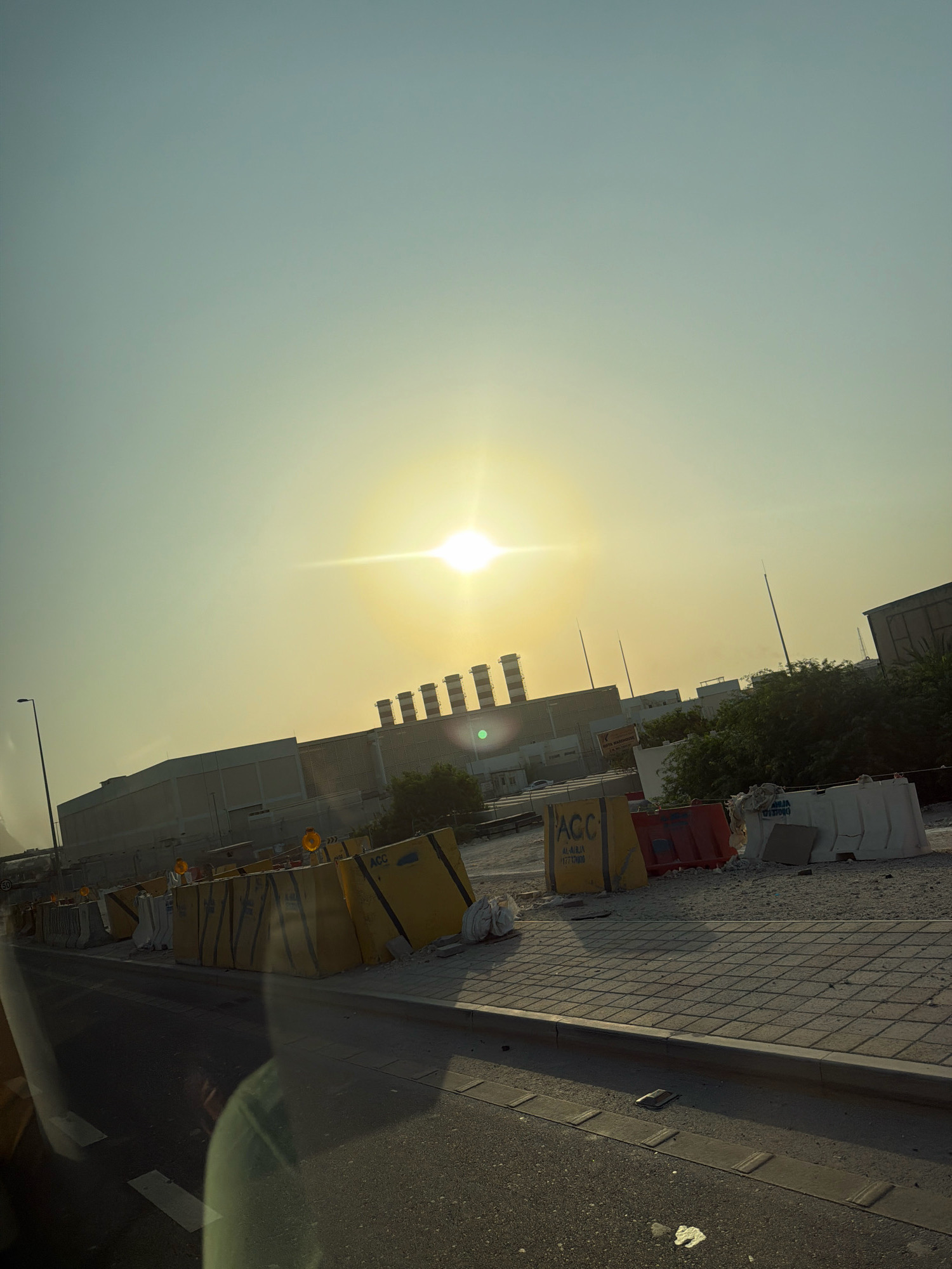 The image captures a sunlit scene in Riffa, Bahrain. The sun is a bright, lens-flared orb high in the pale sky, casting a golden hue over the environment. In the middle ground, a large industrial-looking building complex with distinctive smokestacks is visible. In front of the complex, construction barriers, primarily yellow and white concrete blocks, line the side of a road. Some of these barriers have signage, including one with the text "ACC AL-RIJA". A sidewalk with a checkered pattern runs alongside the road, separated by a curb. The road itself is paved and appears to be a thoroughfare, with faint lane markings. The overall impression is one of ongoing development or industrial activity under a bright sky.