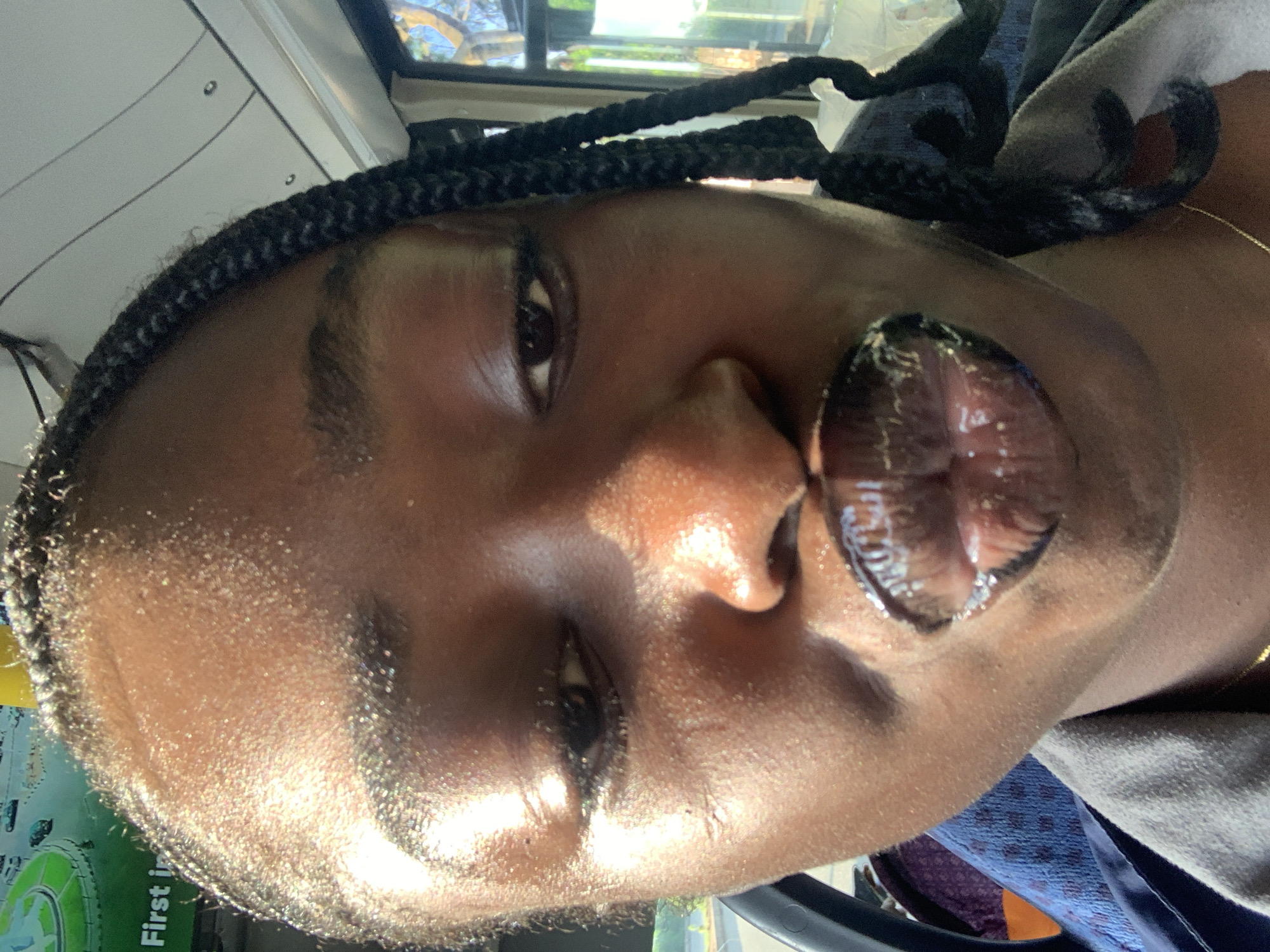 A close-up, selfie-style photograph captures a young Black woman with braided hair. Her face is the primary focus, with her lips pursed and coated in a dark, glossy lipstick. Sunlight glints off her forehead and cheeks, indicating a bright, outdoor environment. She appears to be seated, possibly in a vehicle, as suggested by a glimpse of a blue patterned fabric and a grey garment. The background is blurred, with hints of windows and what looks like foliage outside, implying a public transit setting or a location with natural surroundings. There is no discernible indication of the city of Rafin Par, Nigeria.