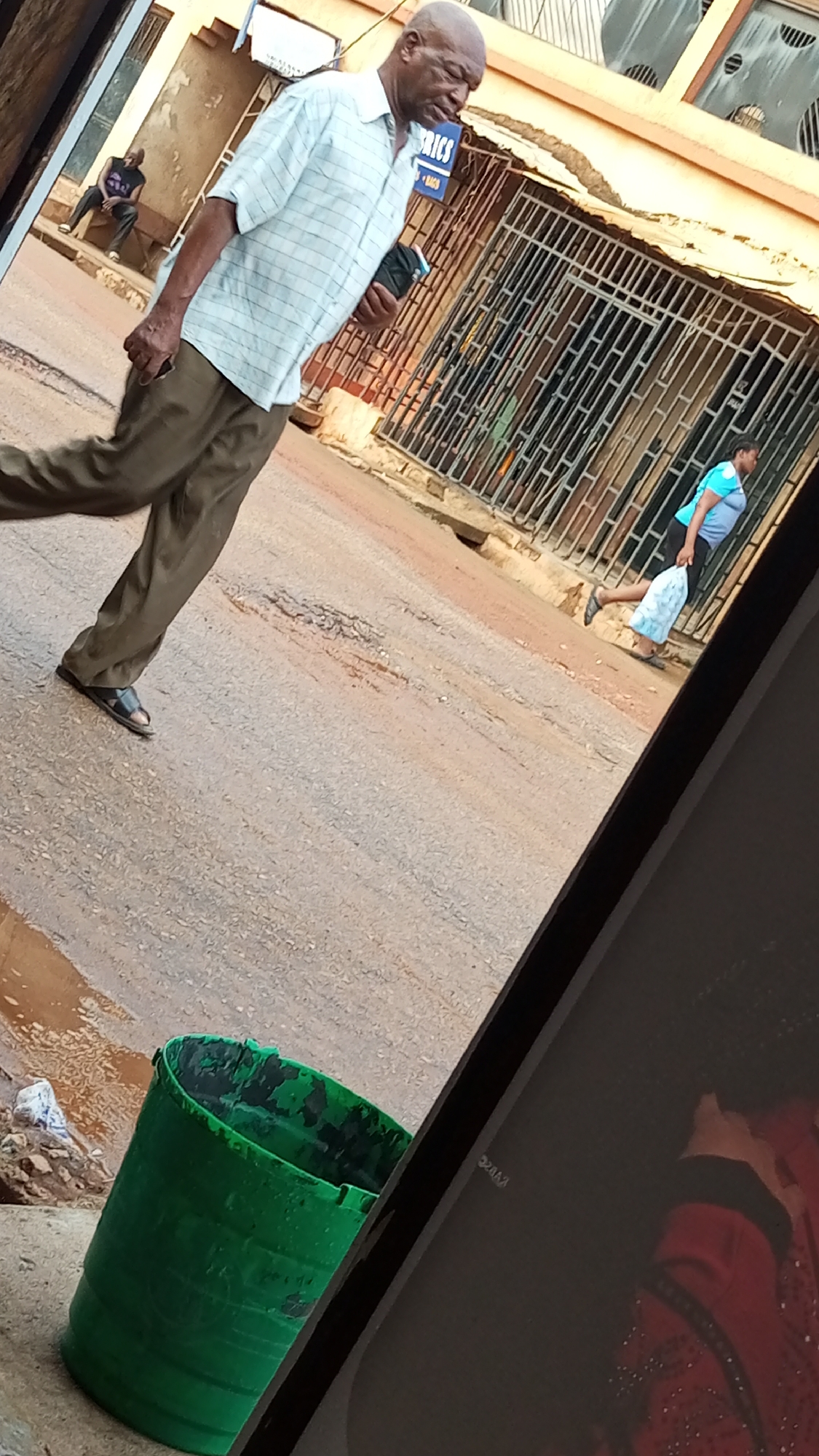 The image shows a street scene in Sabon Tasha, Nigeria. An older, bald African man wearing a light-colored checkered shirt and brown pants walks towards the left of the frame, holding what appears to be a phone or small object in his hand. He is wearing sandals. The street surface is asphalt with visible wear and patches. A green plastic bucket or container sits near the lower left corner. In the background, a building with a metal security gate is visible. A person wearing a blue shirt and dark pants walks away from the viewer toward the gate. Another individual sits on a bench near a building in the distant background.