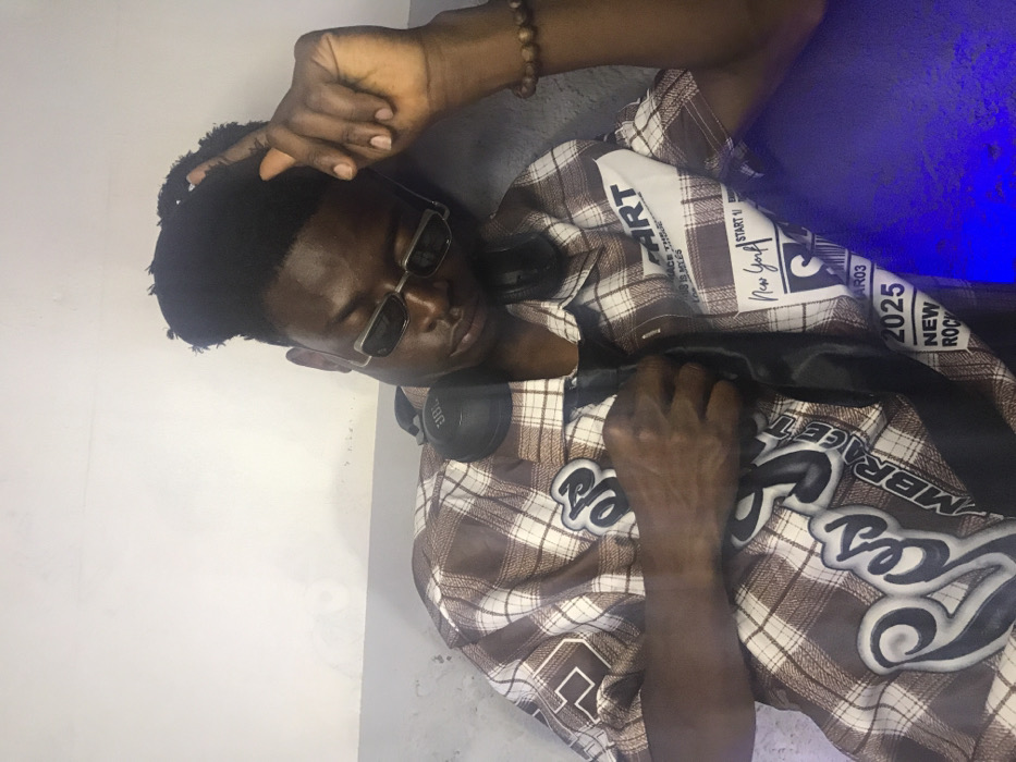 A young Black male is depicted in an indoor setting in Kirana, Nigeria, positioned against a light-colored wall with a darker gray lower section. He wears a short-sleeved brown and white plaid button-up shirt featuring multiple stitched patches, including "ROSES," "EMBRACE," "START," "New York," "2025," and "NEW ROCK." A black satin tie is visible beneath his shirt collar. He wears rectangular dark sunglasses and black JBL over-ear headphones resting around his neck. A brown beaded bracelet is on his right wrist. His dark hair is styled with volume on top. His head is tilted slightly to the right, and his eyes are closed or directed downward. His left hand is raised, fingers slightly parted, near the left side of his head. His right hand is positioned on his chest, lightly gripping a section of his shirt and tie. A distinct blue light source illuminates the lower right portion of the frame, casting a blue glow on his shirt.