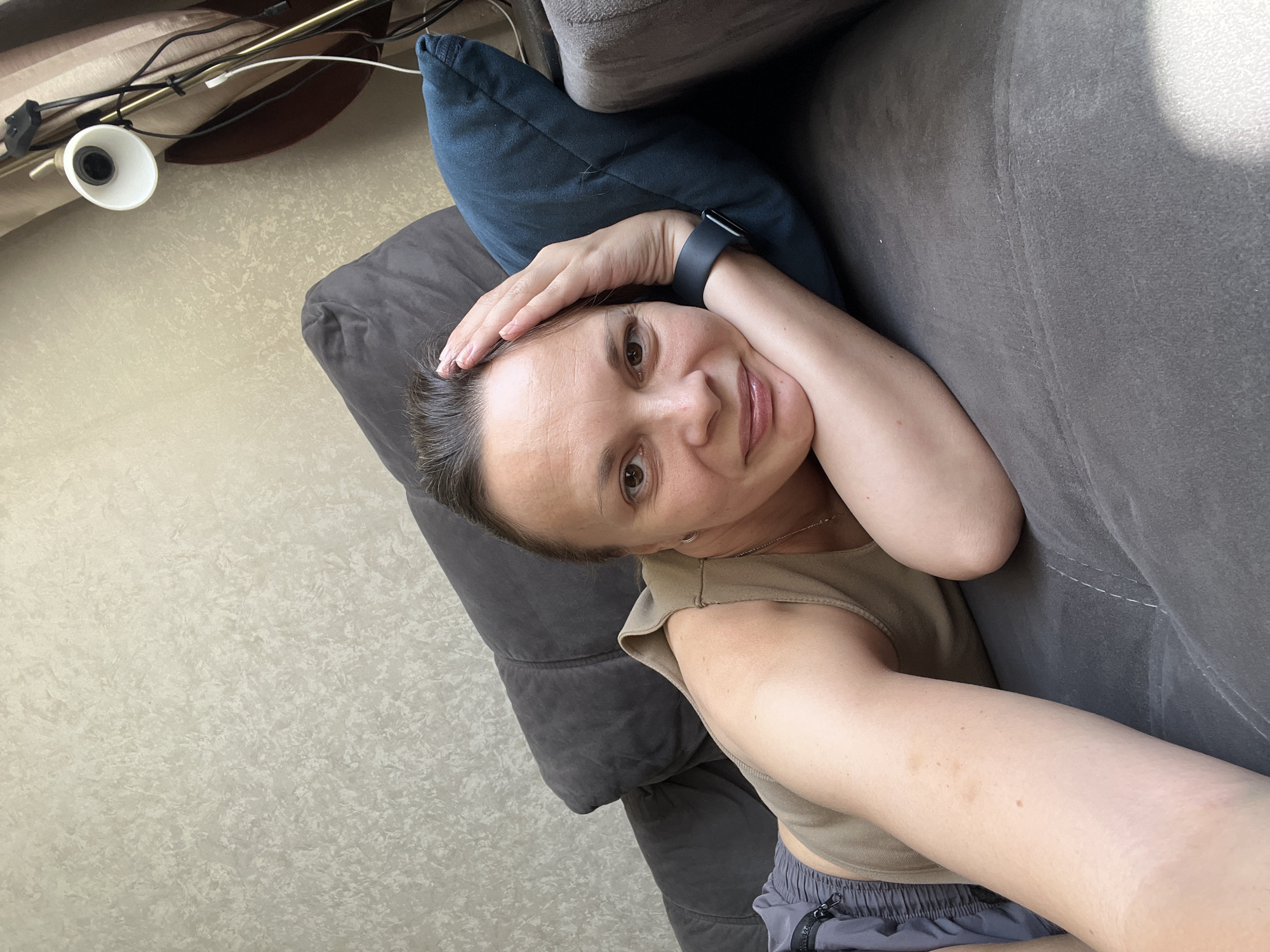 A woman is lying on a sofa, resting her head on a blue pillow. She is looking directly at the camera and smiling. She is wearing a beige tank top and grey shorts. A smartwatch is visible on her left wrist. The background shows a textured wall and a floor lamp. The image does not provide any information about the location in the city of Tolyatti, Russia.