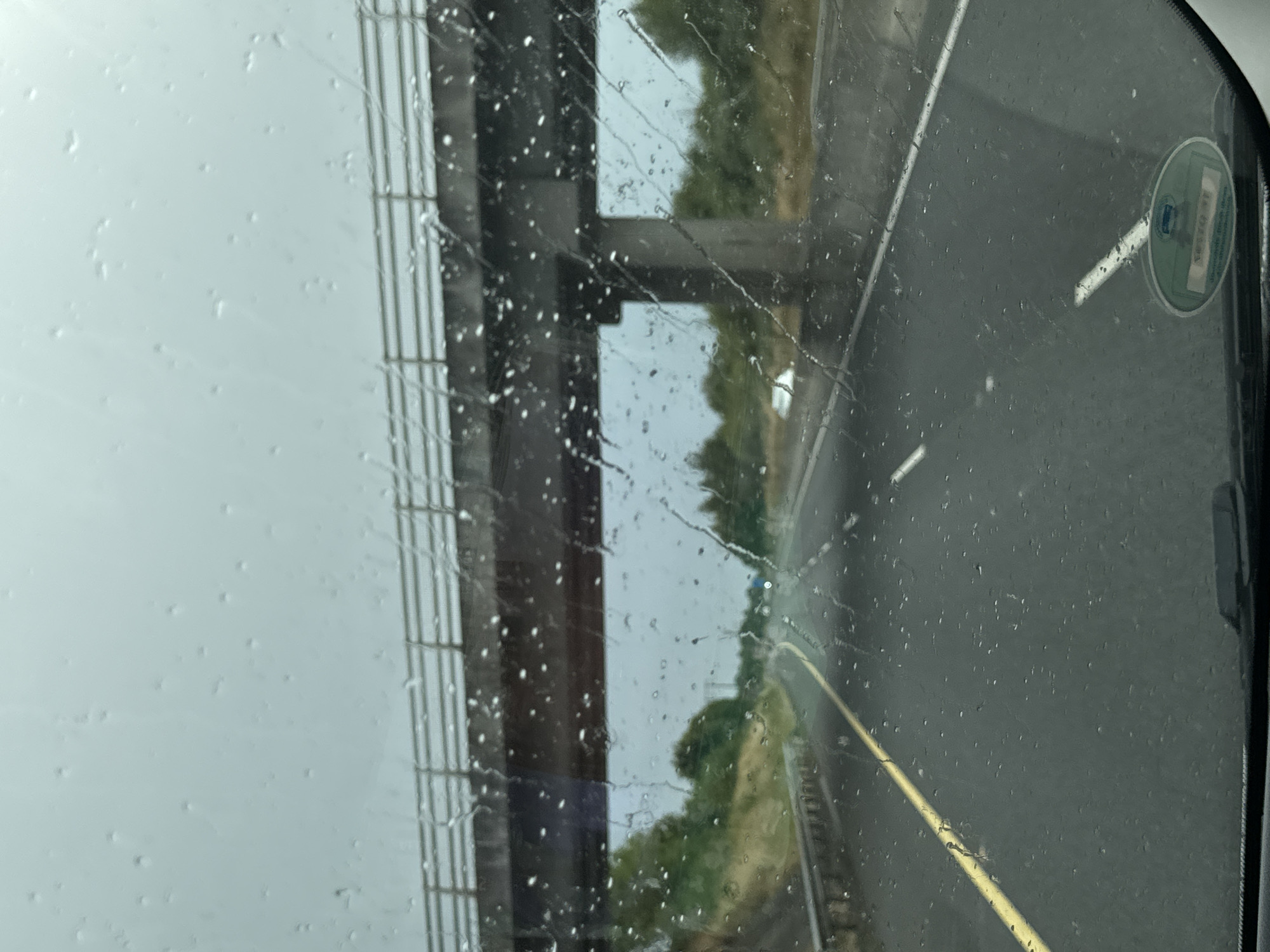 An image taken from inside a vehicle on a highway in Ireland shows a highway overpass in the distance with rain drops on the windshield obstructing the view. The road ahead is wet and has a yellow line marking the lane. Green vegetation and trees line the sides of the highway. The sky is overcast and grey.