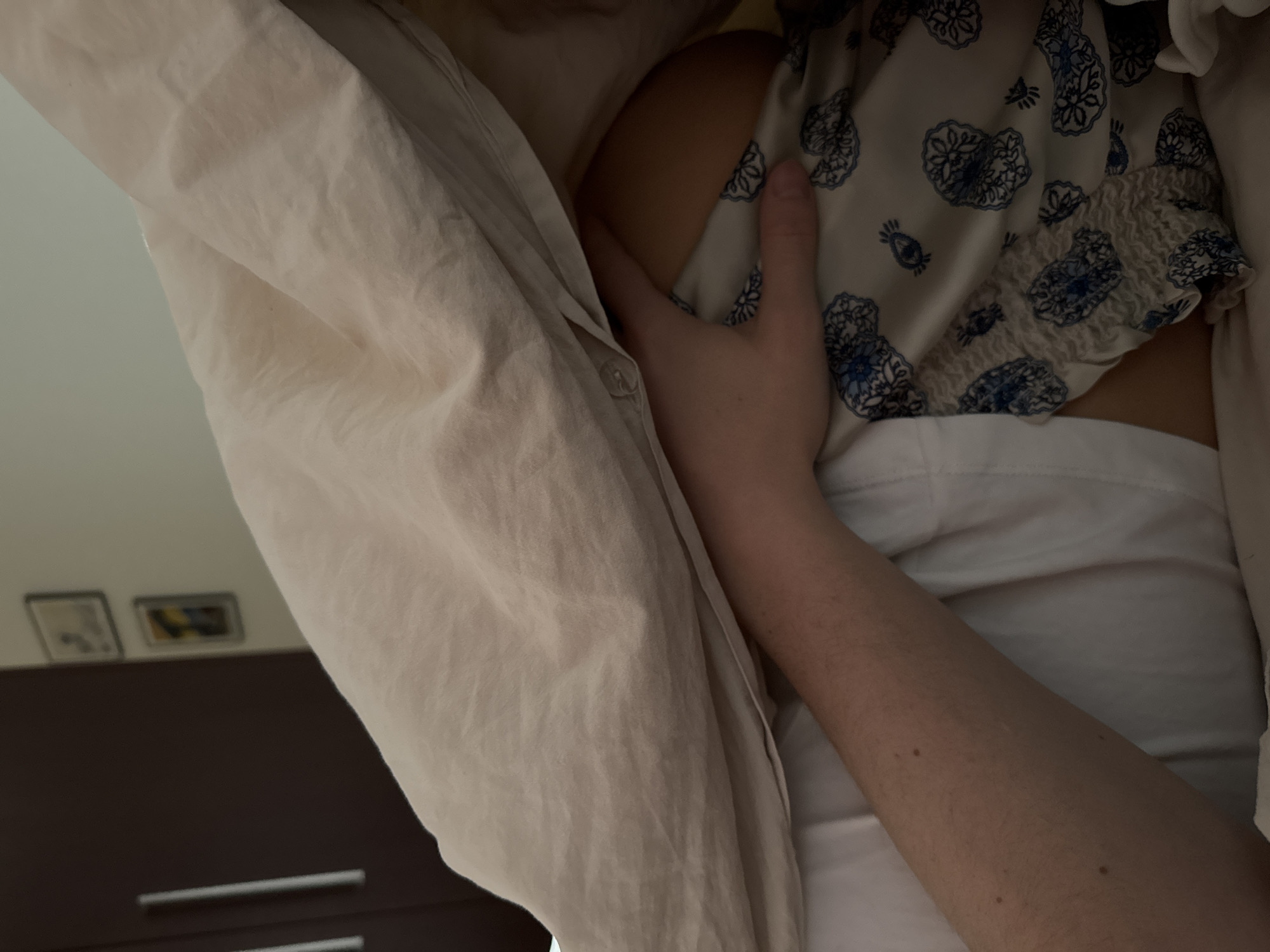 The image shows a person lying in bed, possibly in a bedroom setting. The person is partially covered by a white linen sheet, with a portion of their upper body visible. They are wearing a light-colored garment with blue floral patterns and shirring. A hand is positioned on the person's upper arm or shoulder. In the background, parts of a dark brown dresser are visible, with two framed pictures above it. The location is unknown.