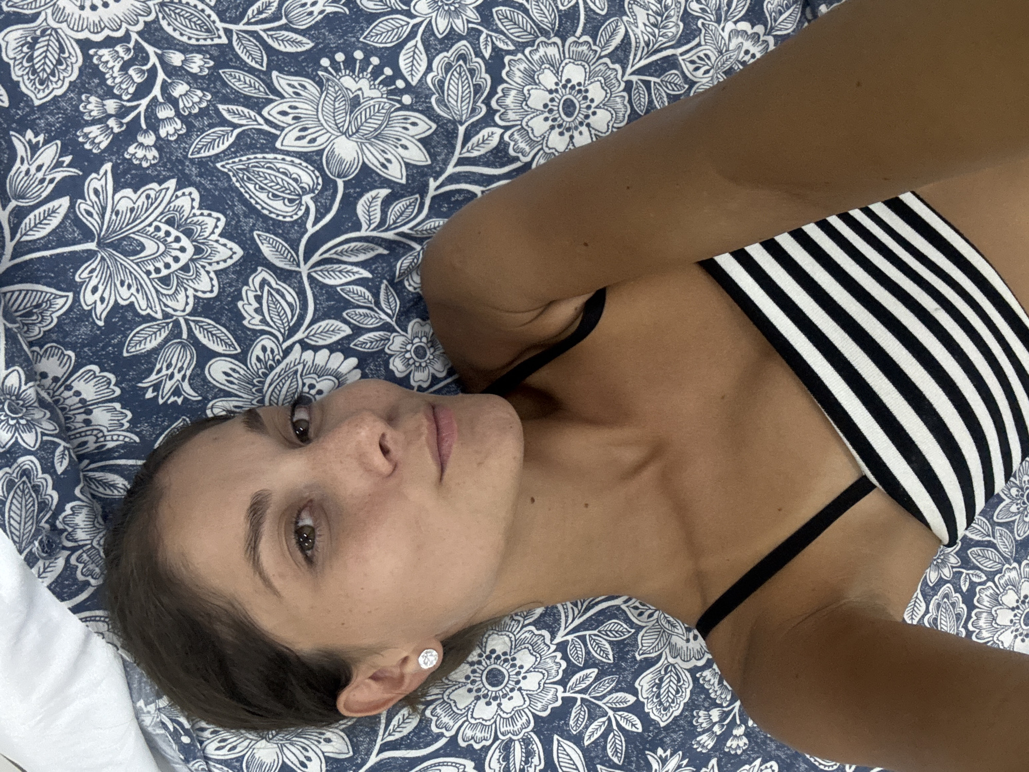 The image features a young woman lying on her back, looking directly at the camera. She has brown hair, dark eyes, and a neutral, calm expression. Freckles are visible on her nose and cheeks. She is wearing a black and white striped bikini top with thin black straps, and a small, round, stud earring is visible in her left ear. Her skin appears tanned.
The woman is positioned on a surface covered by a dark blue fabric adorned with a white floral and leaf pattern, likely bedding or a blanket. A portion of a plain white sheet is visible in the bottom-left corner. The angle of the shot, with her arm slightly raised and bent, suggests she may be taking a selfie. The setting appears to be indoors, likely in Kuala Lumpur, Malaysia, based on the provided context. There is no visible text in the image.