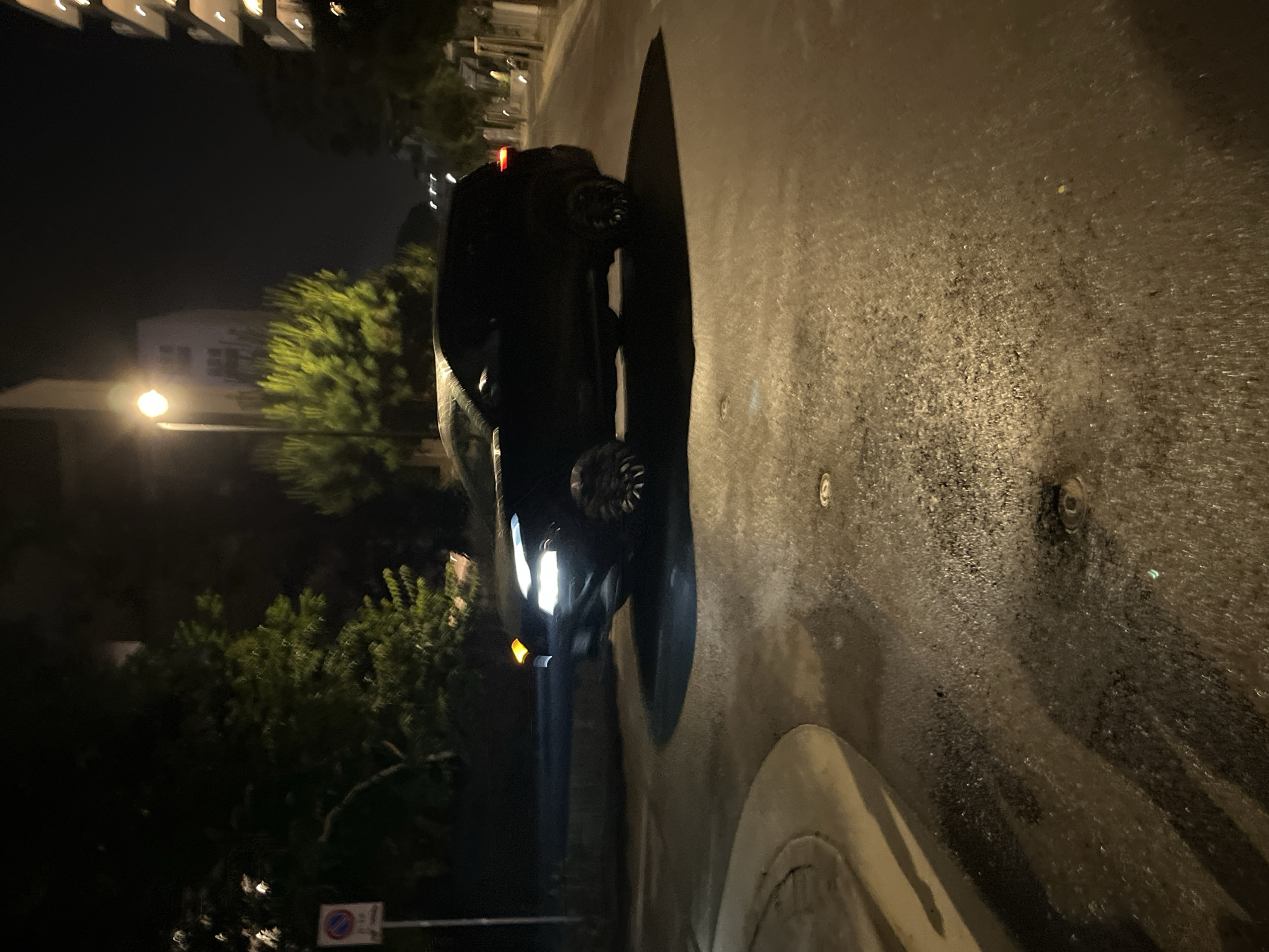 The image depicts a dark and wet street scene at night in Jesolo, Italy. A dark-colored car is parked on the right side of the road, with its front facing the lower portion of the image. Streetlights illuminate the area, casting reflections on the wet road surface. Trees and buildings can be seen in the background, and a street sign is visible on the left side. The overall environment suggests an urban or suburban setting during nighttime.