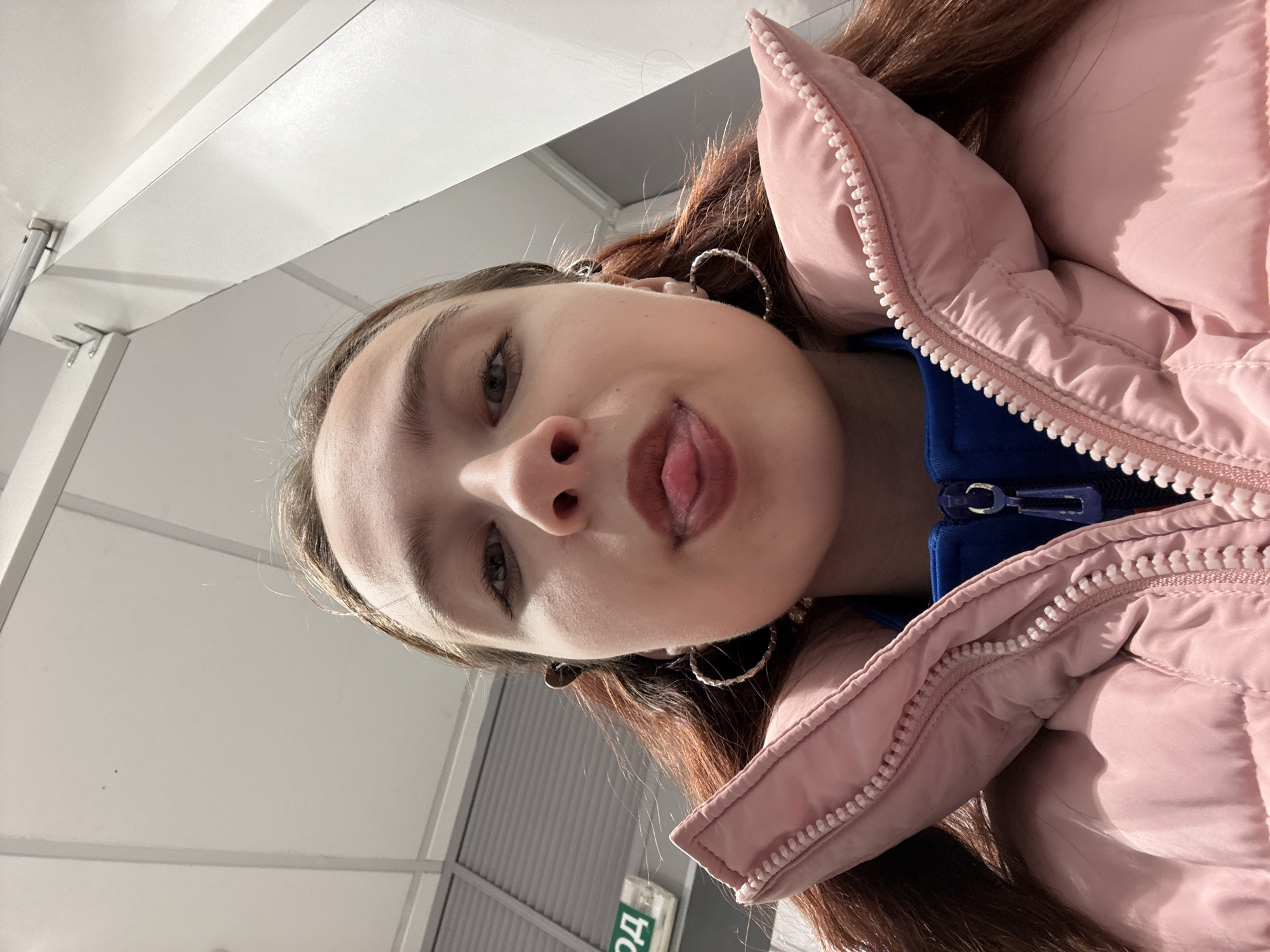 A close-up, angled view features a person with light skin, brown hair, and fair eyebrows, wearing hoop earrings. They are sticking out their tongue. The person is wearing a blue top under a pink puffer jacket. The background reveals a commercial-style ceiling with metal supports and a sign with Cyrillic characters visible. The location appears to be indoors, possibly a commercial or public space.