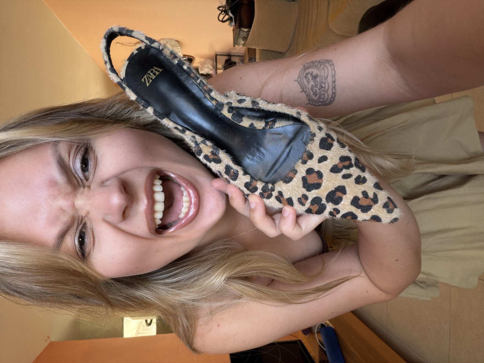 A woman with blonde hair is holding a leopard-print shoe, with the ZARA logo visible, close to her face while smiling widely. She has a tattoo on her arm. The location is not discernible.